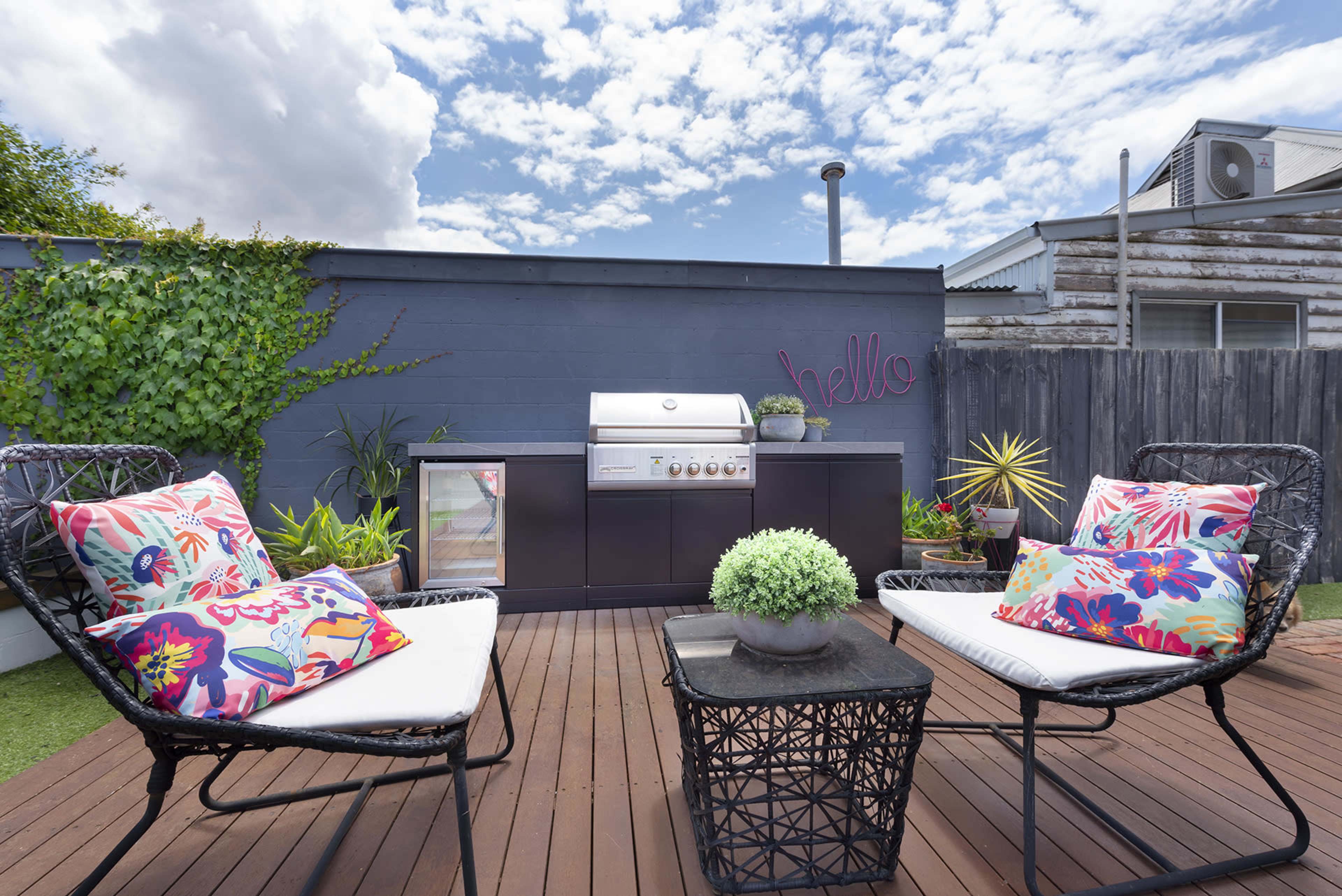 A backyard patio featuring a barbecue grill, potted plants, and two chairs with colorful cushions on a wooden deck.