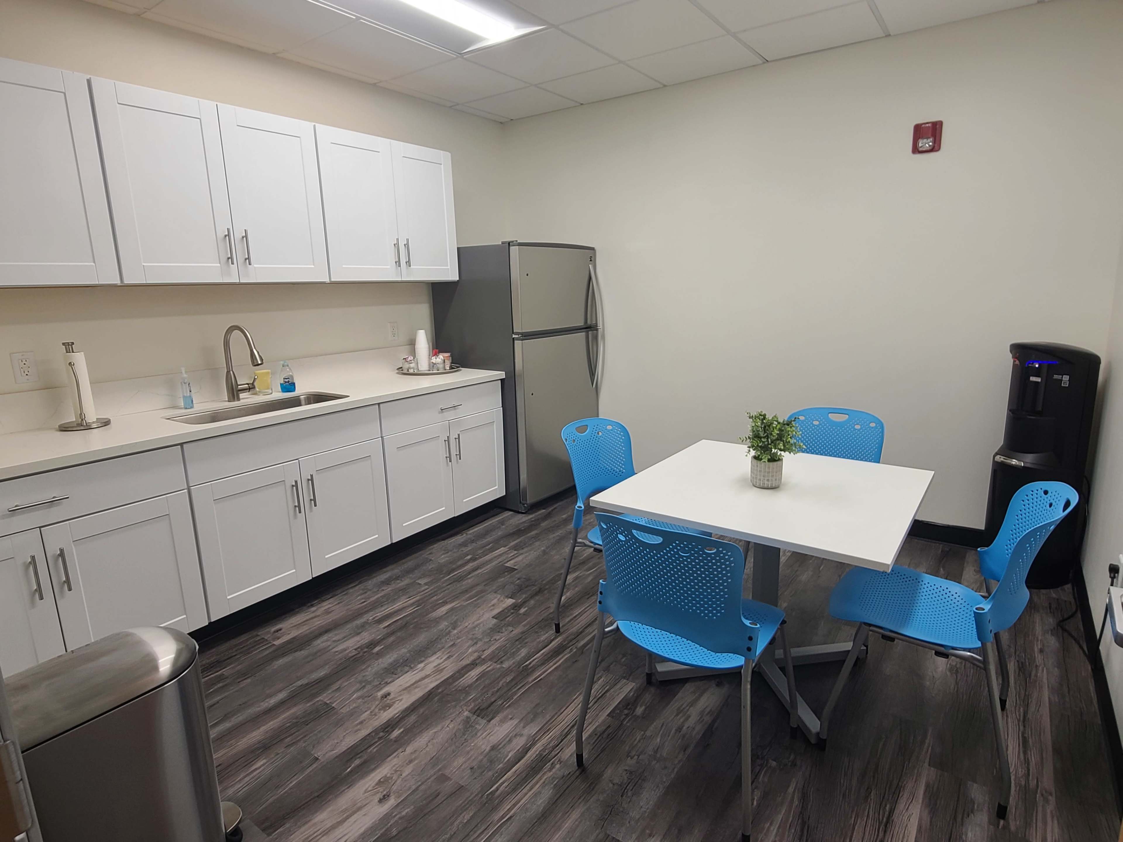 A clean break room featuring a table with four blue chairs, a refrigerator, and a sink against white cabinets, with a small plant on the table.