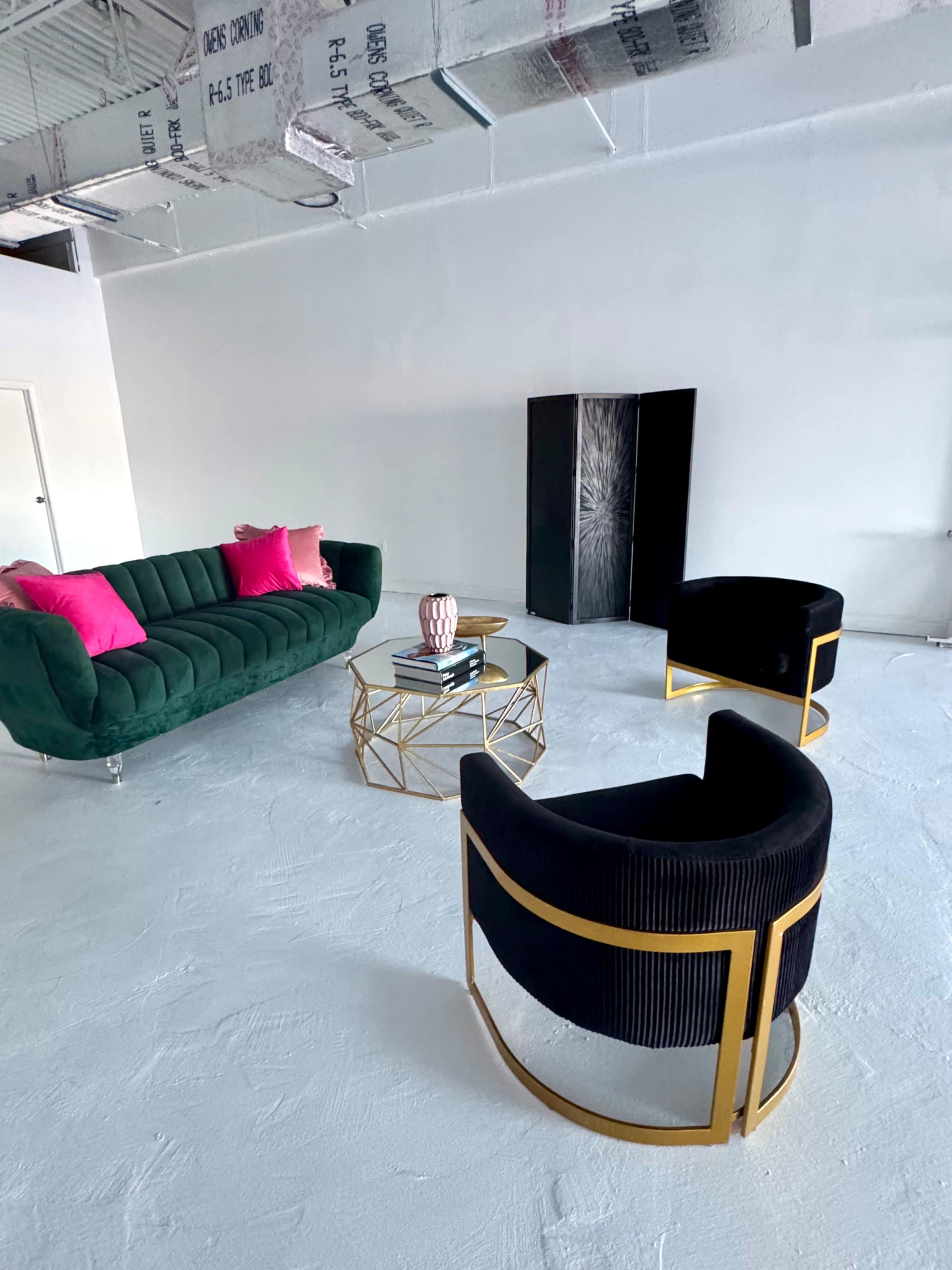 The image shows a modern, minimalist room featuring a green velvet sofa, two black armchairs, a glass and gold coffee table, and a tall black cabinet.