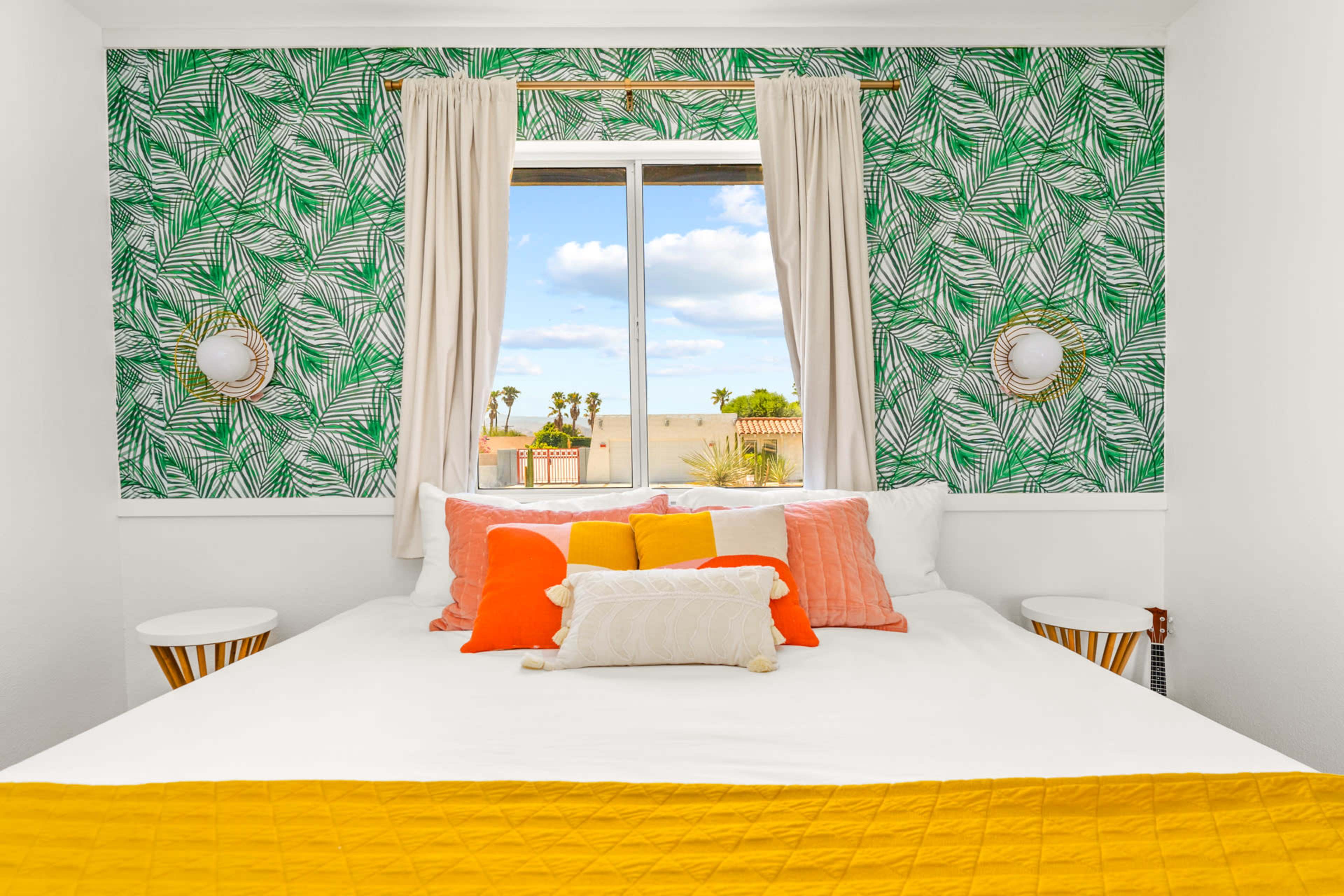 A bright bedroom features a bed adorned with colorful pillows against a leafy green wallpaper and a window showcasing a view of palm trees.