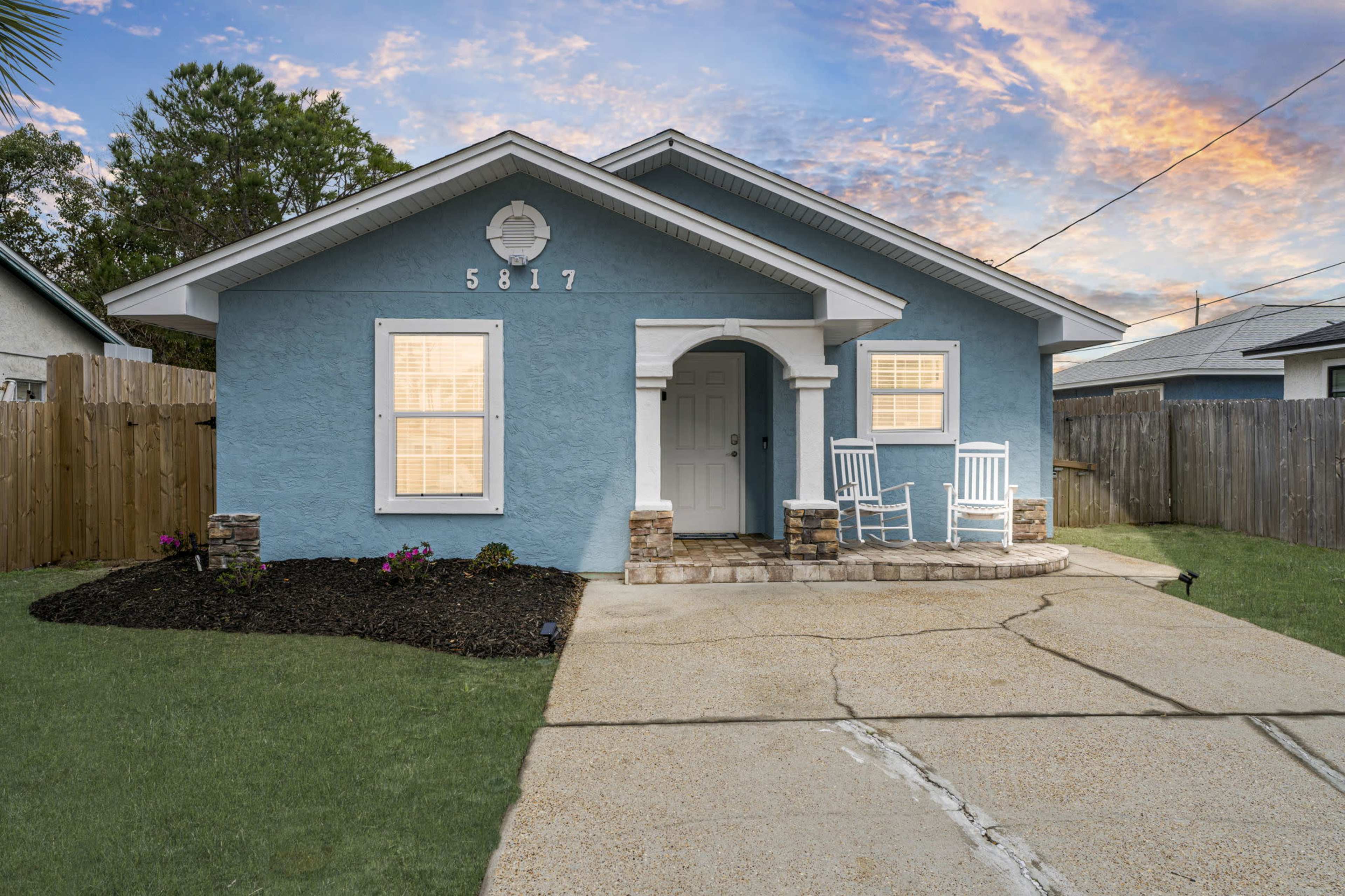 The image features a light blue house with a front porch, two rocking chairs, and a concrete driveway.