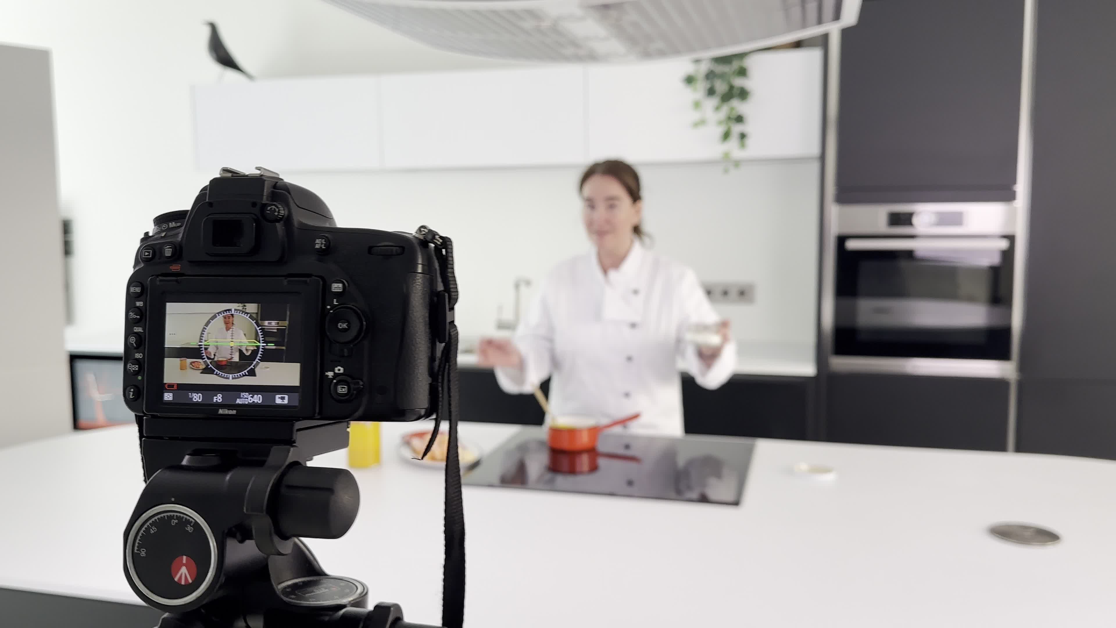 A camera on a tripod is focused on a woman in a chef's jacket gesturing while standing behind a kitchen island with a pot on the stove.