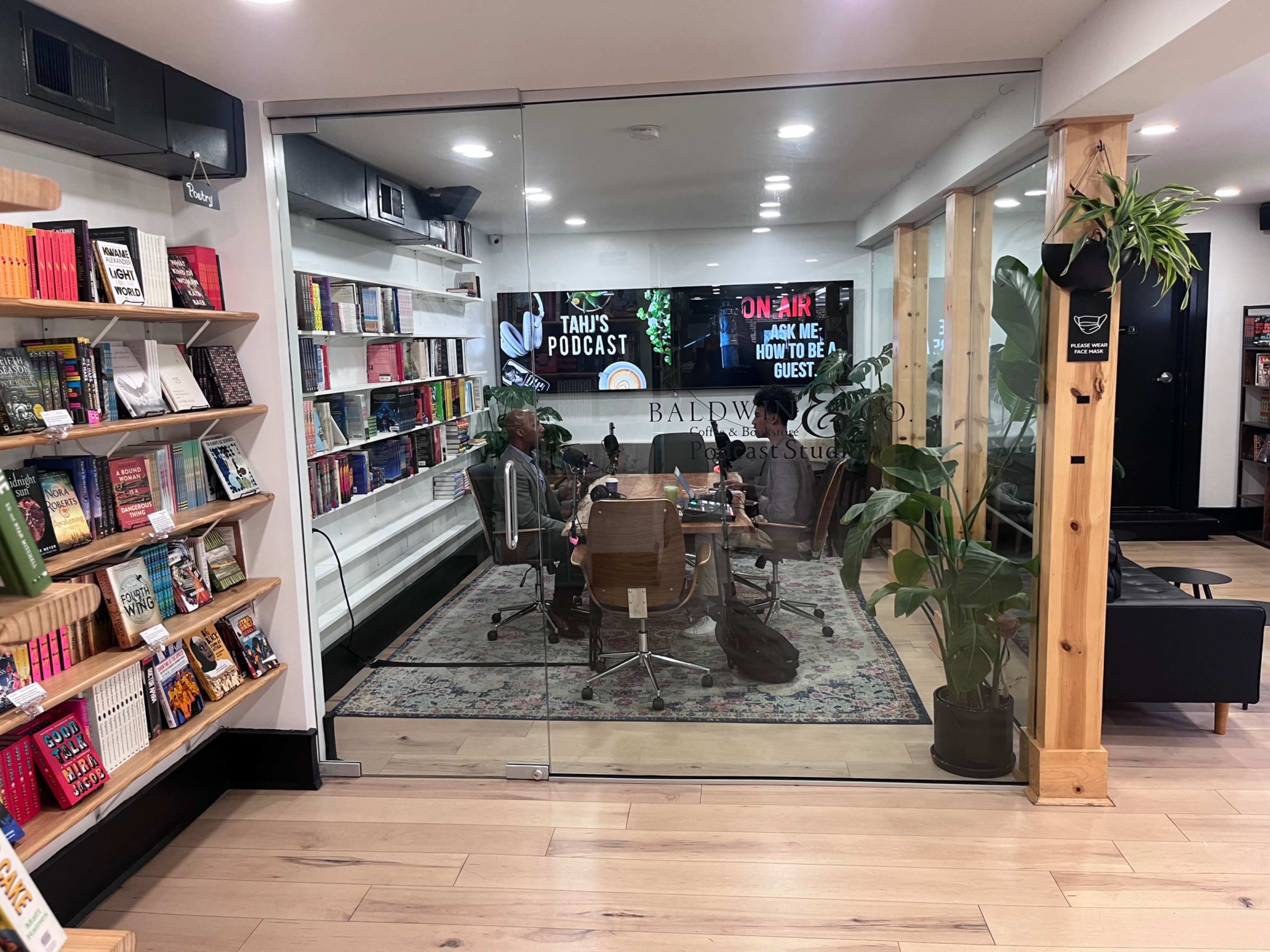 The image shows a podcast studio with a glass entrance, featuring a round table and chairs surrounded by bookshelves and plants.