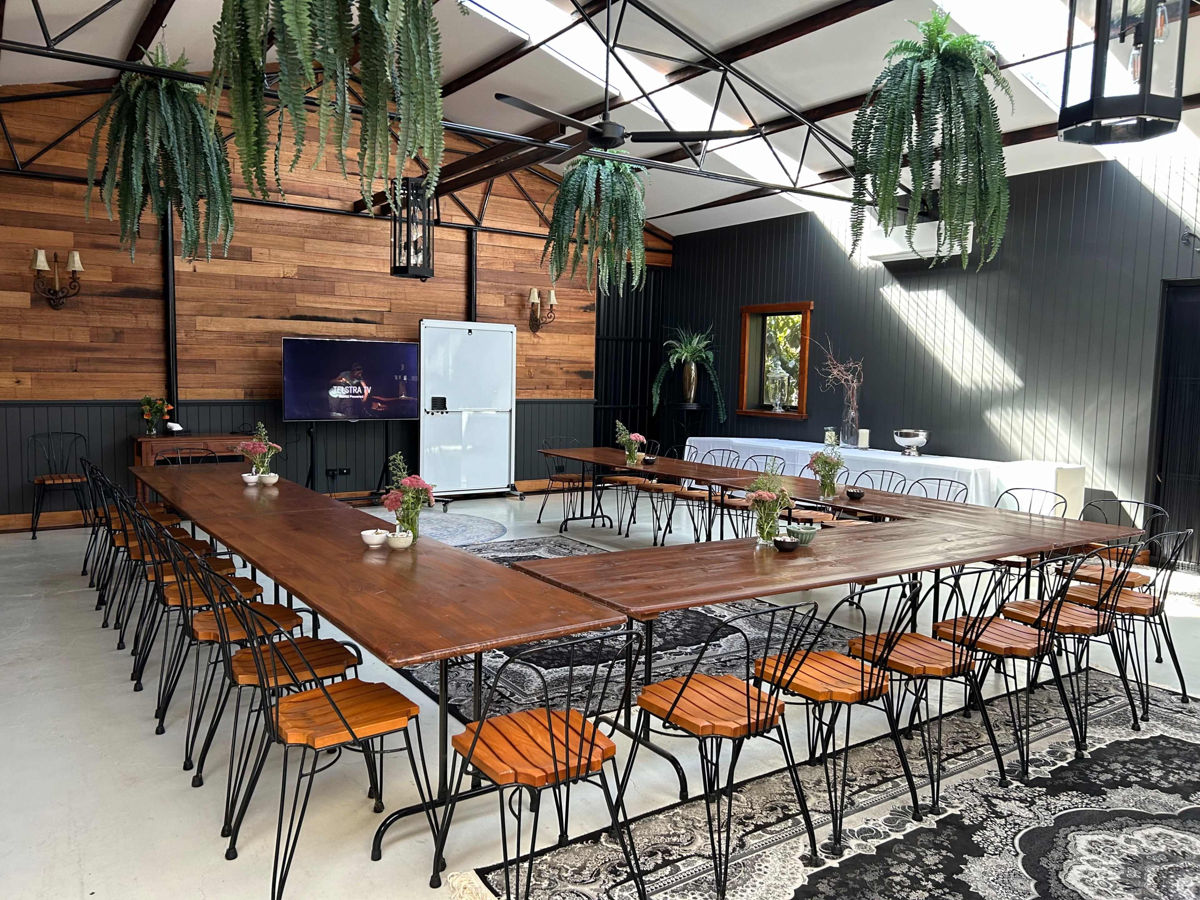 A long wooden table surrounded by metal chairs fills a bright, airy meeting room with greenery and decorative elements.