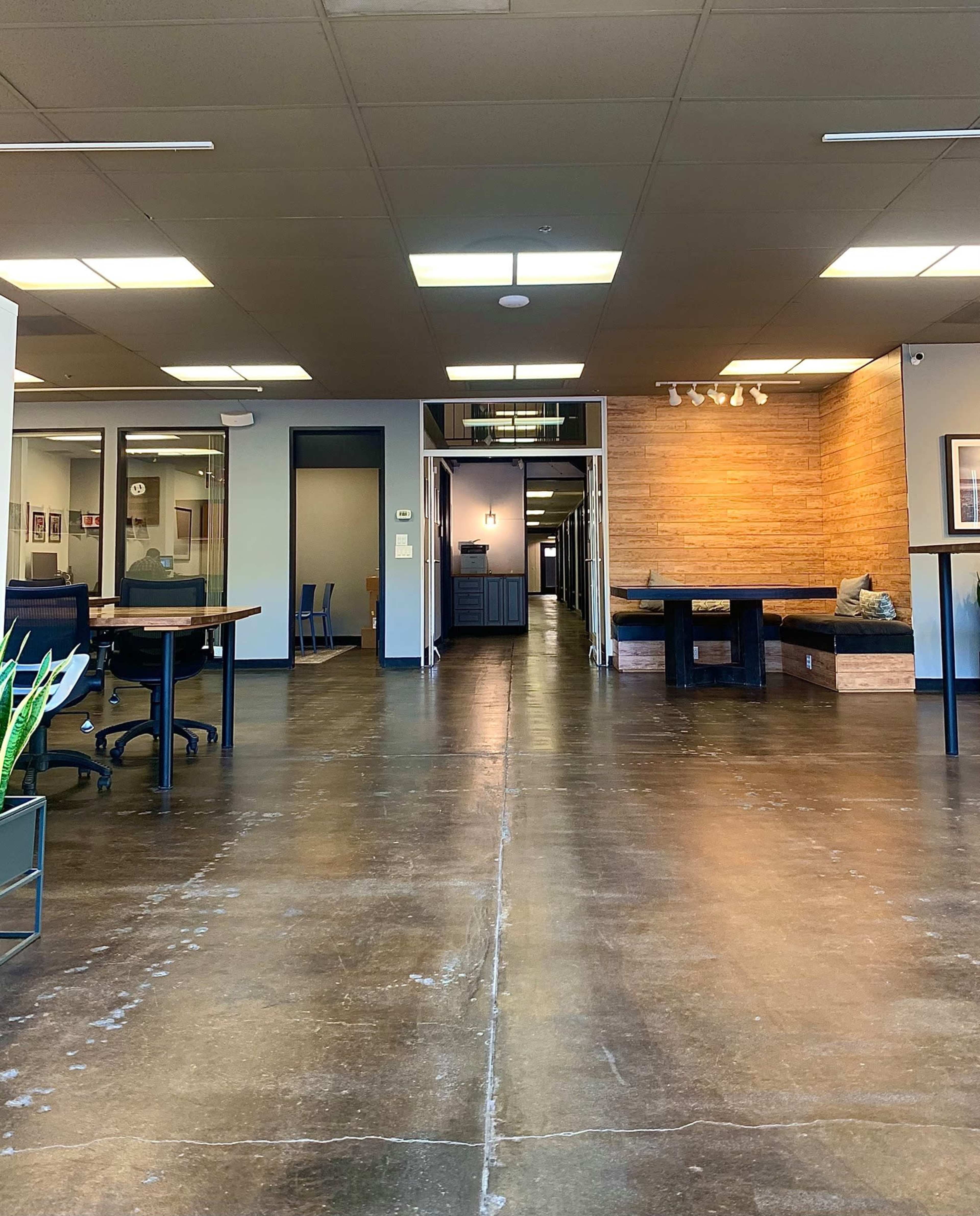 An open office space with a polished concrete floor, featuring several workstations on the left and a hallway leading to a wooden-paneled area in the background.