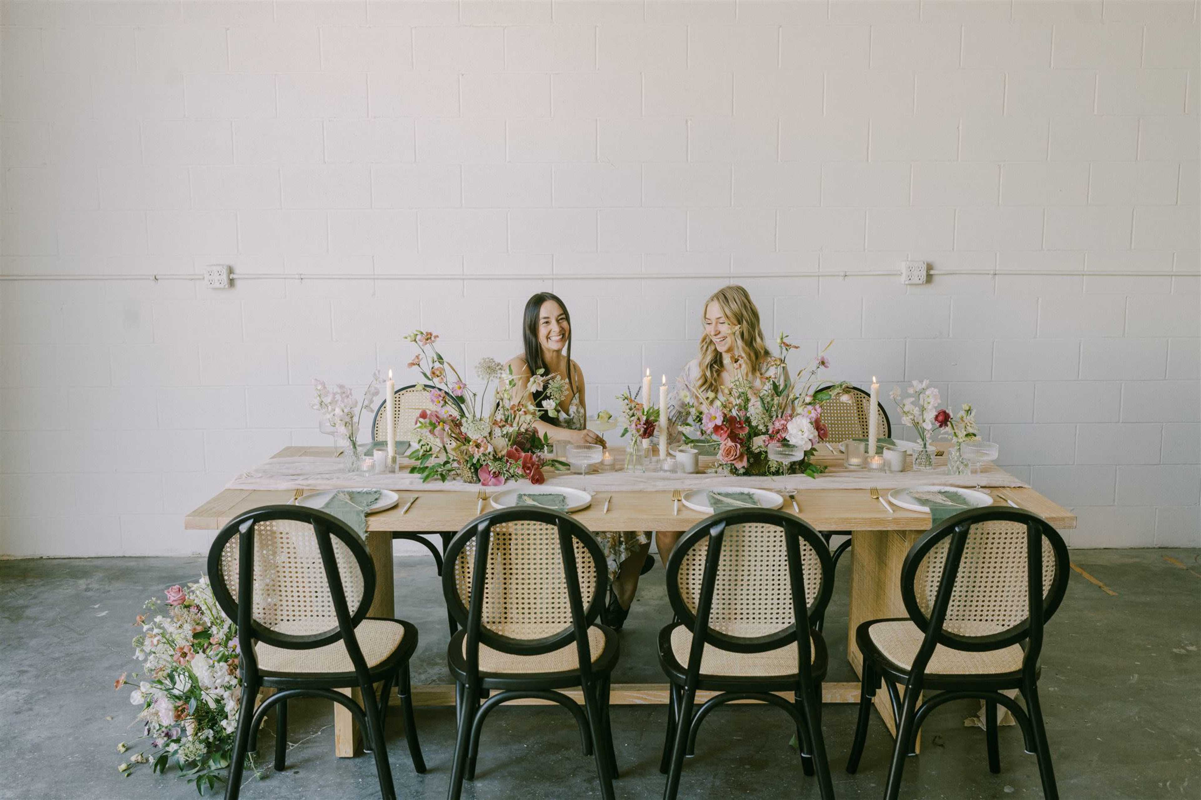 Two women sit at a long wooden table decorated with floral arrangements and candles in a minimalist room.