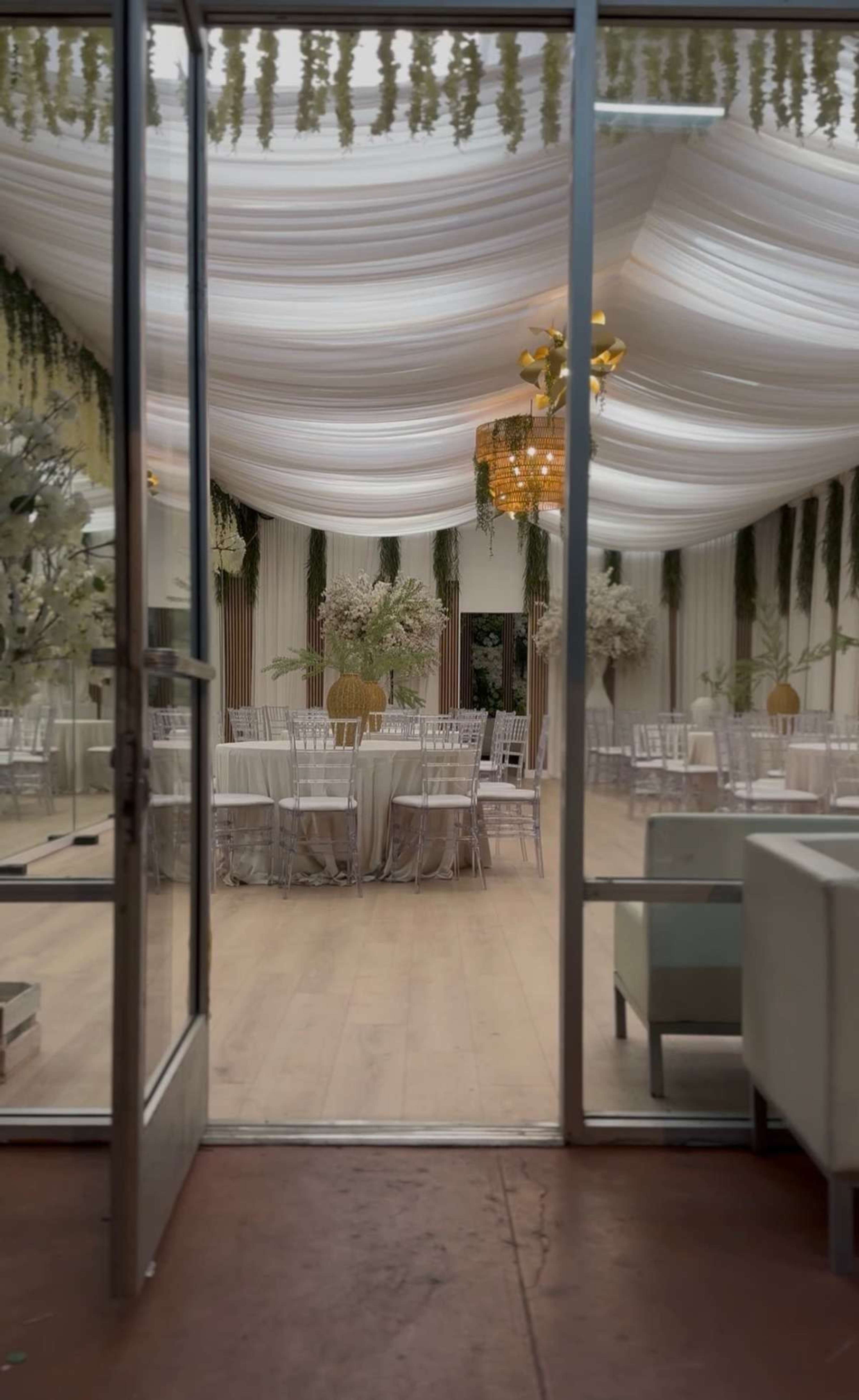 The image shows a well-decorated indoor event space with draped ceilings, tables set for an occasion, and floral arrangements throughout.