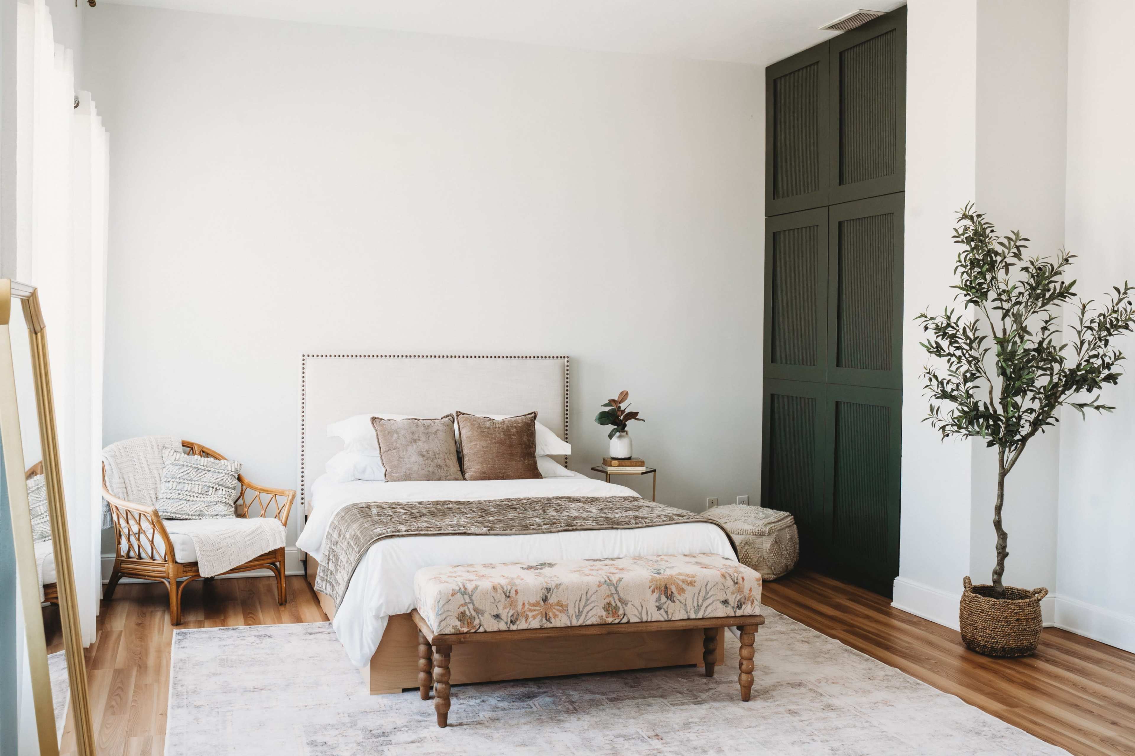 The image shows a bright bedroom featuring a bed with decorative pillows, a wooden bench, a plant, and a chair, all against a white wall.