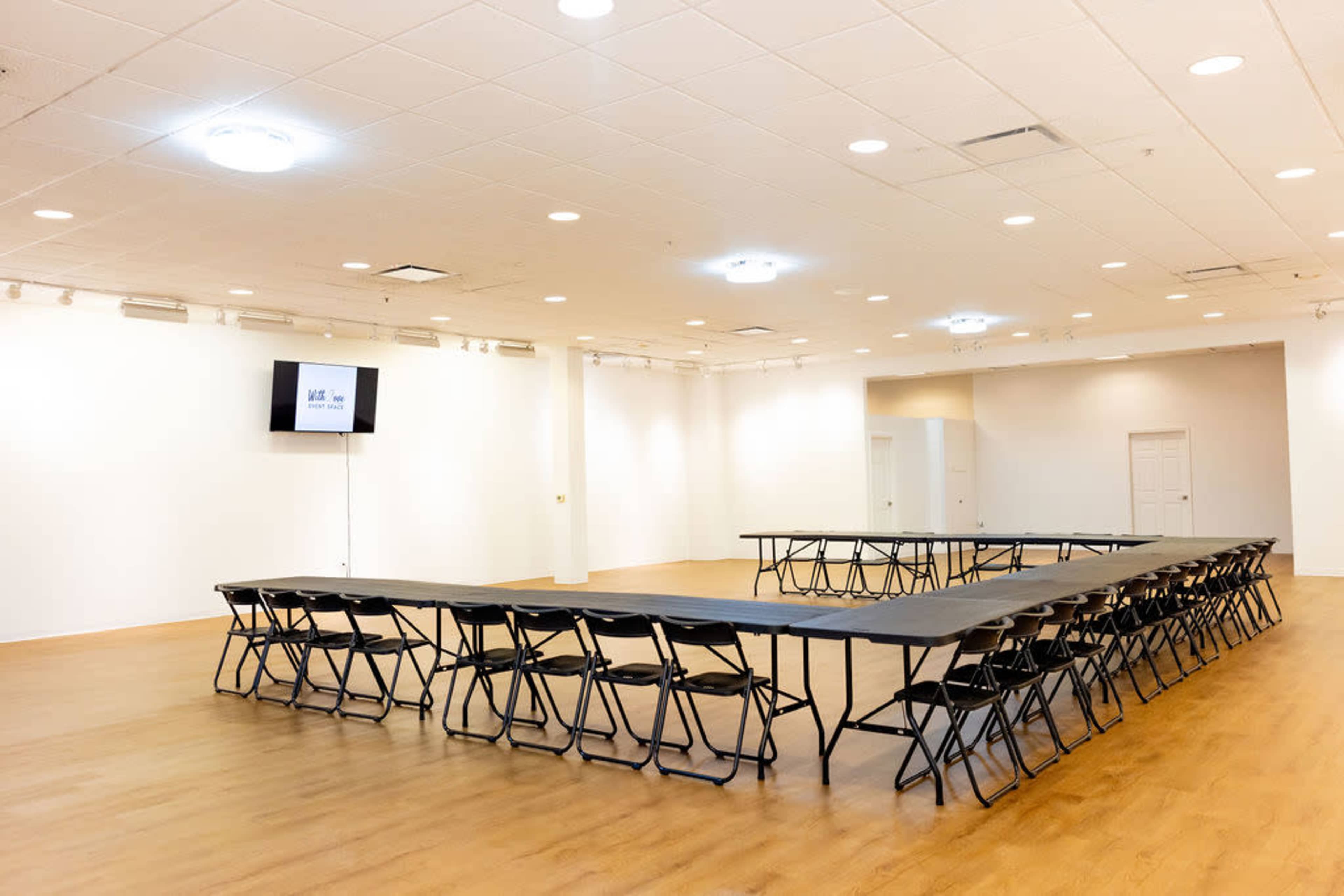 The image shows a spacious meeting room with a large U-shaped arrangement of black folding chairs around a long table, and a screen mounted on the wall.