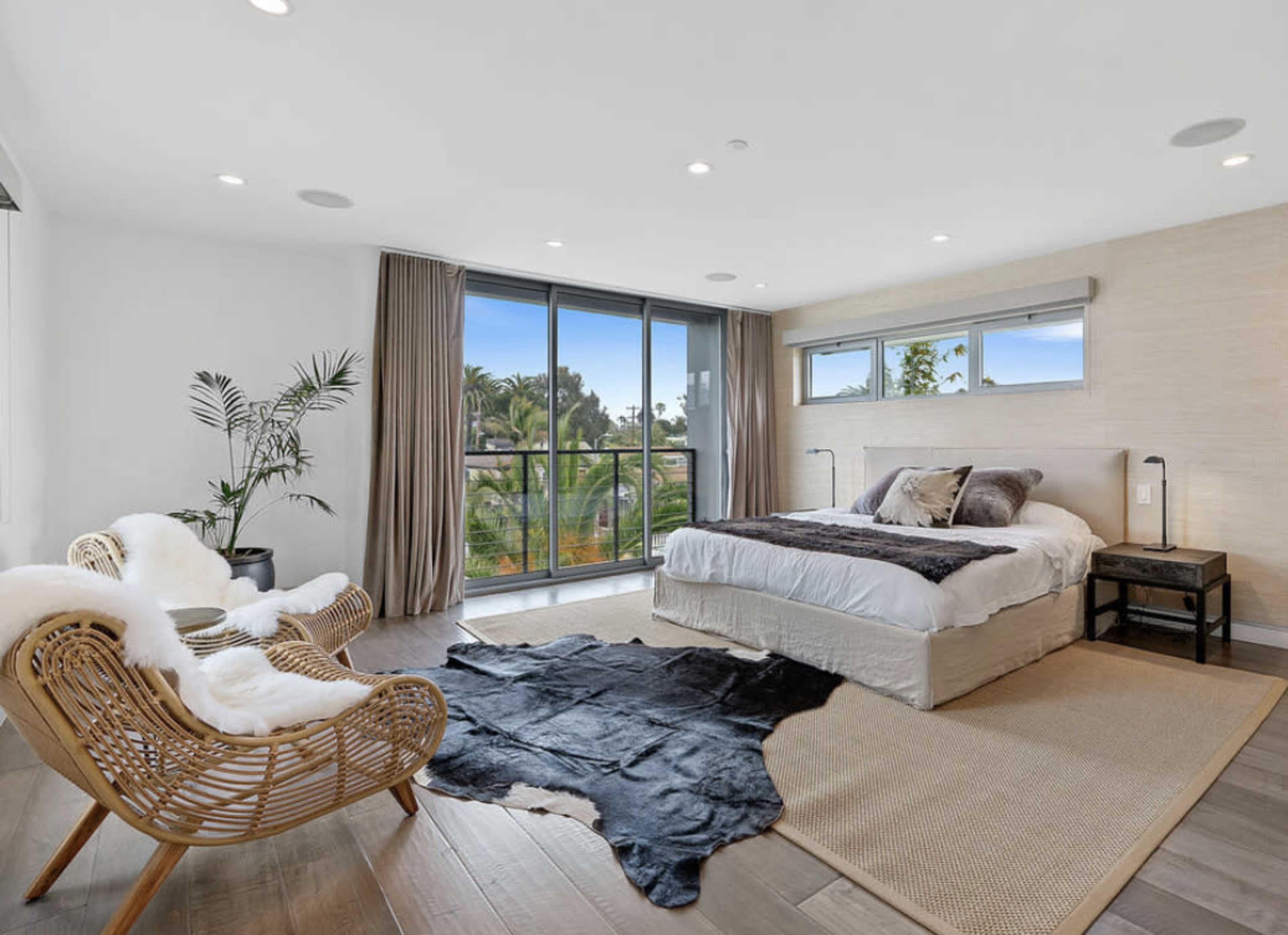 A spacious bedroom features a large bed, two armchairs, a rug, and a view of greenery through large windows.