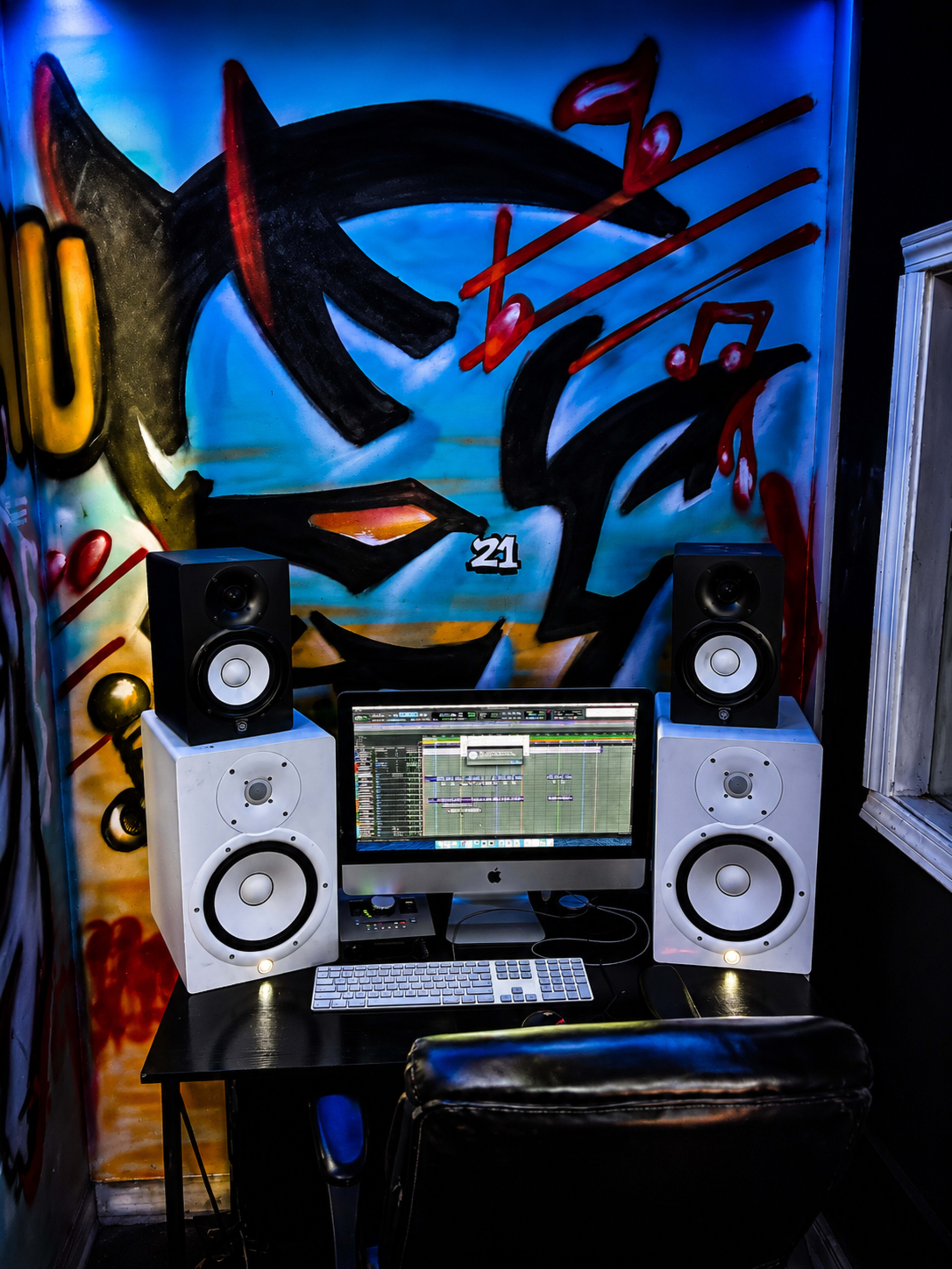 A music production setup features a computer monitor, audio speakers, and a keyboard on a desk in front of a colorful graffiti wall.