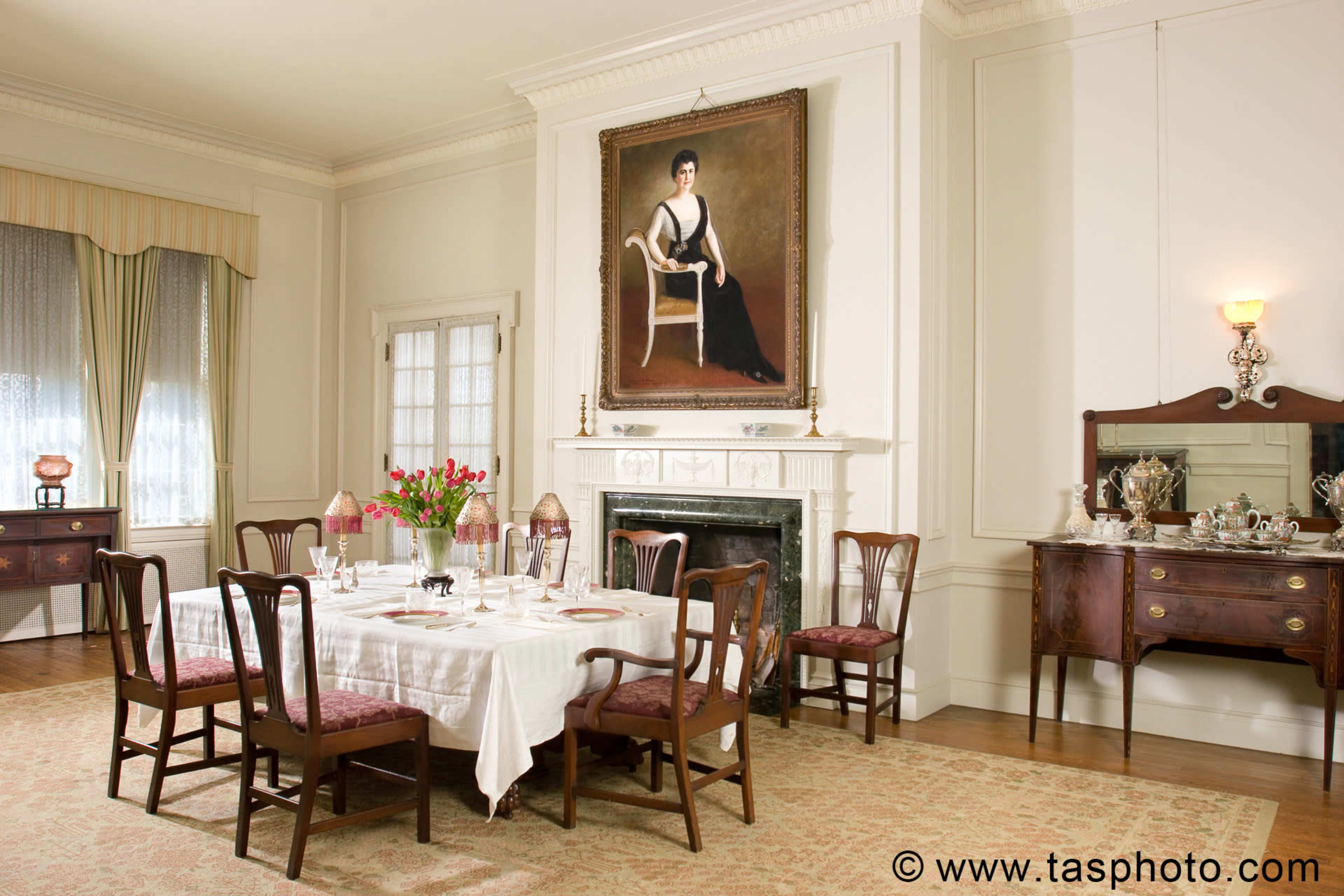 A dining room features a large table set with dishes and flowers, framed by elegant furniture and a prominent portrait on the wall.