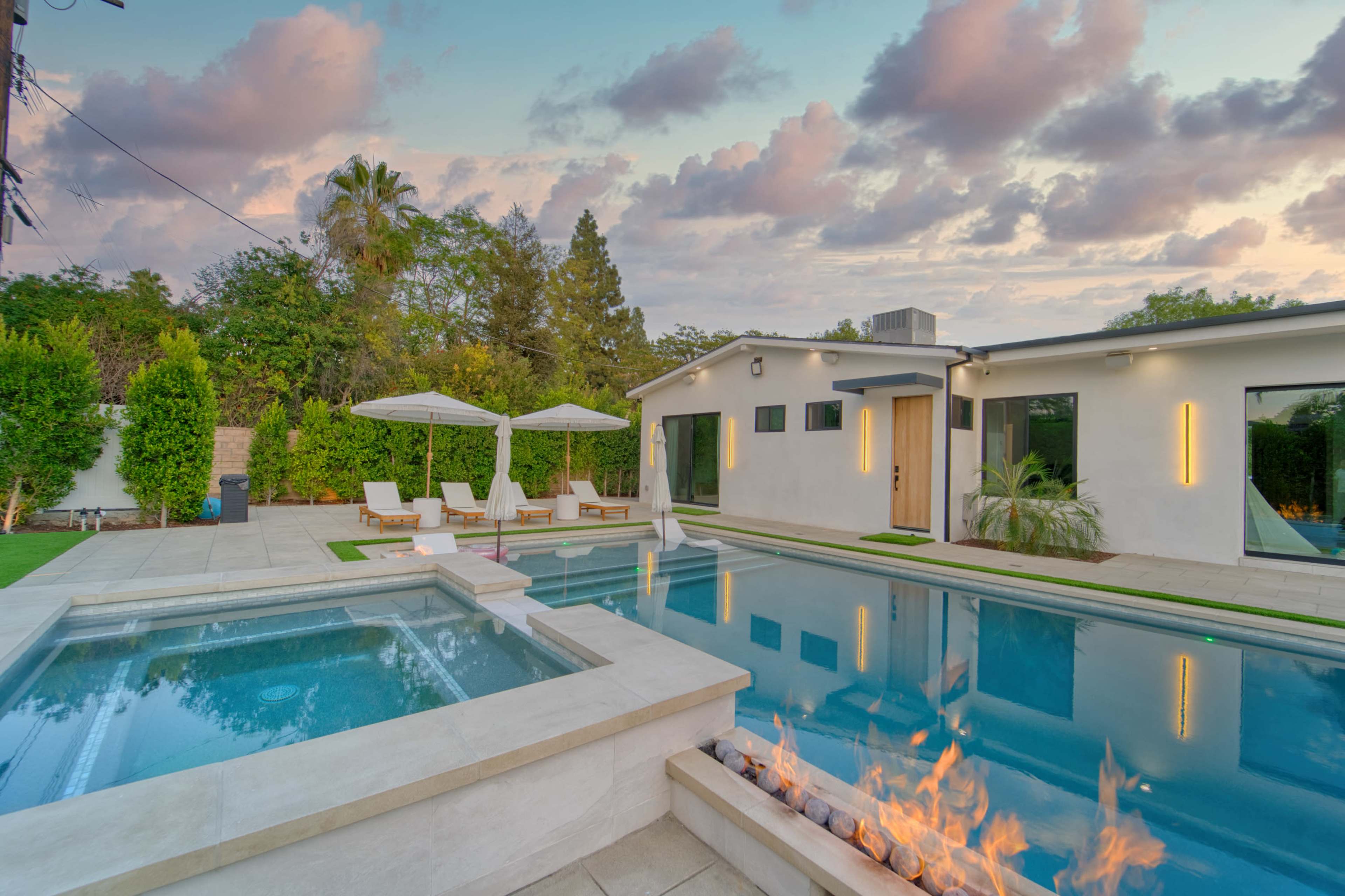 The image shows a modern pool area featuring a sparkling swimming pool, lounge chairs, two umbrellas, and an outdoor fire feature, all set against a backdrop of trees and a colorful sky.