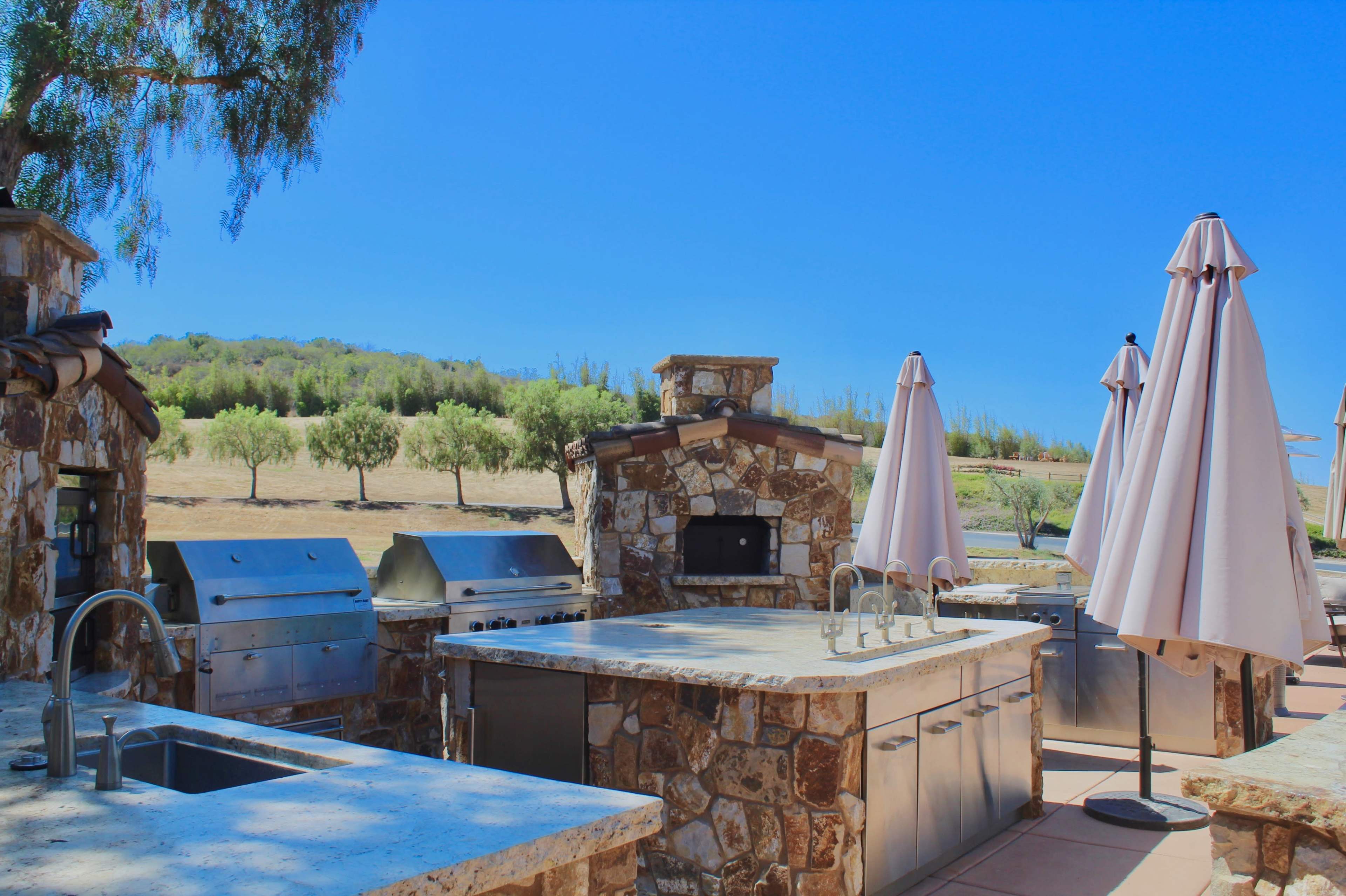 An outdoor kitchen area features stone structures, stainless steel grills, and large umbrellas, set against a hillside with trees.