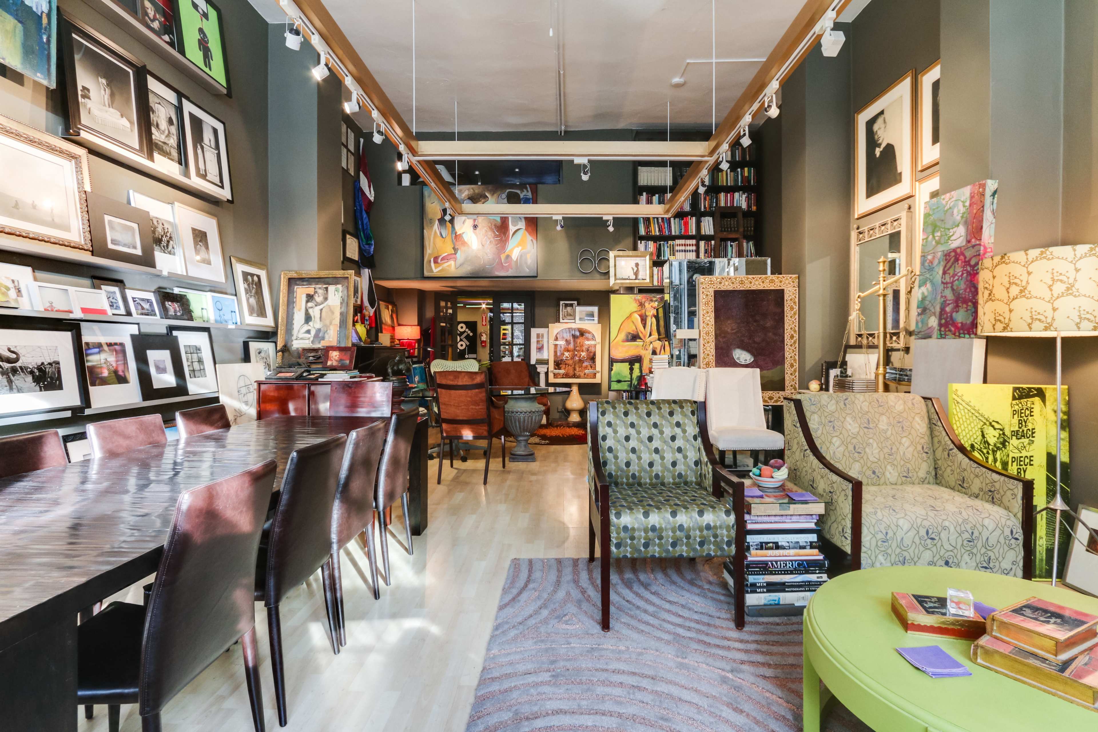 An art-focused interior space featuring a mix of framed photographs and paintings on the walls, various seating arrangements, and a long dining table.