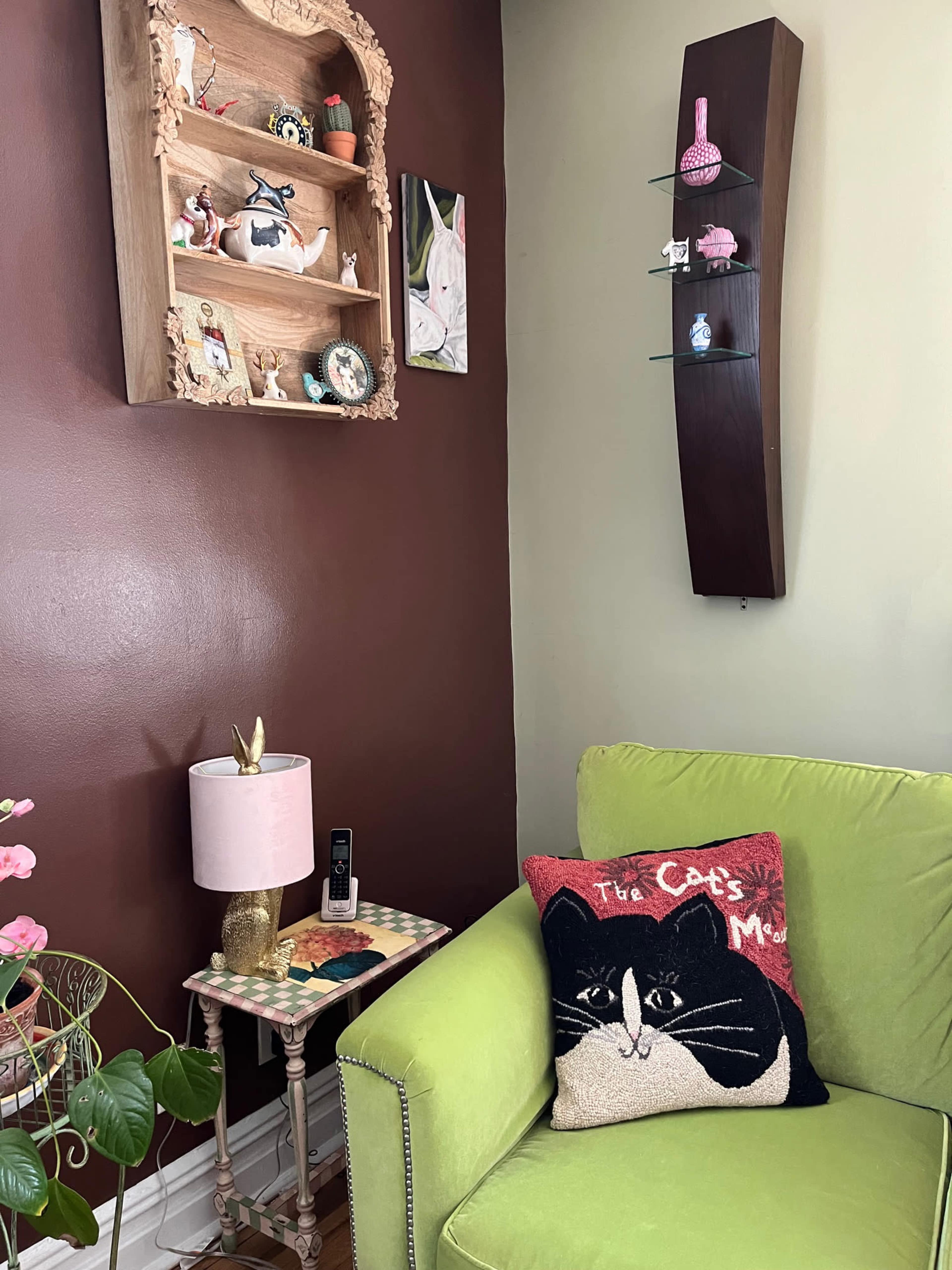 The image shows a cozy corner of a room featuring a green couch, a small side table with a lamp, and a wall shelf displaying decorative items.