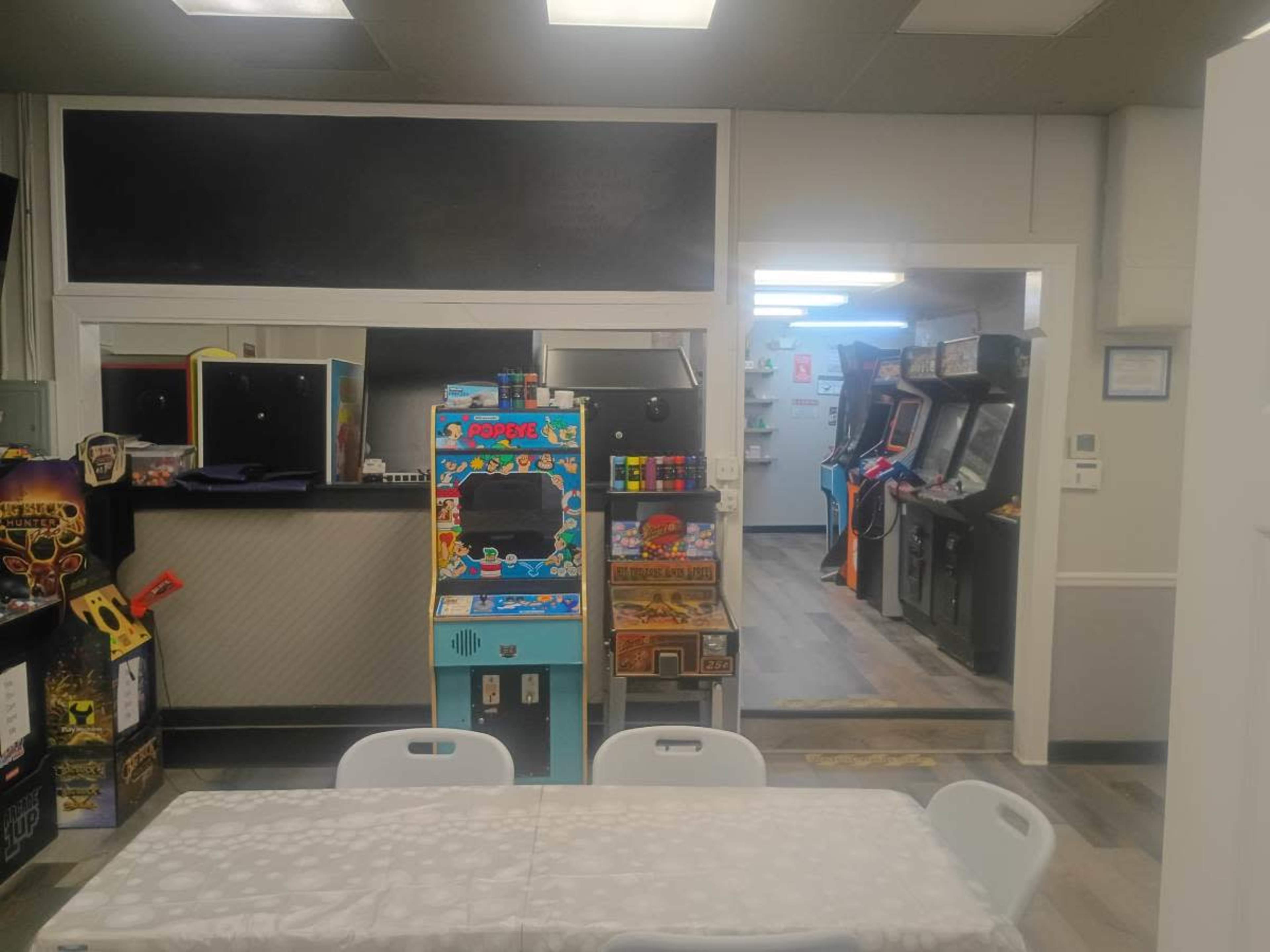 The image shows a game room with various arcade machines arranged along the walls and a table with chairs in the foreground.