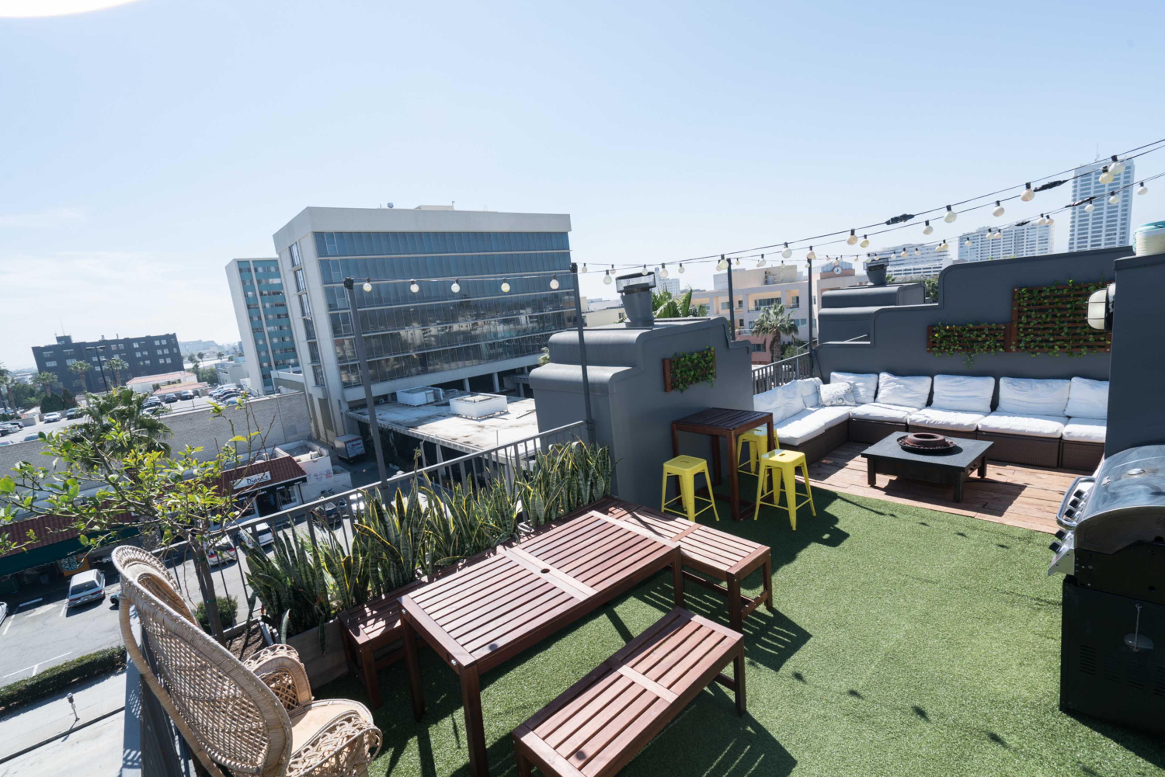 The rooftop features wooden furniture, green plants, and a seating area with a grill against a backdrop of urban buildings and clear blue skies.