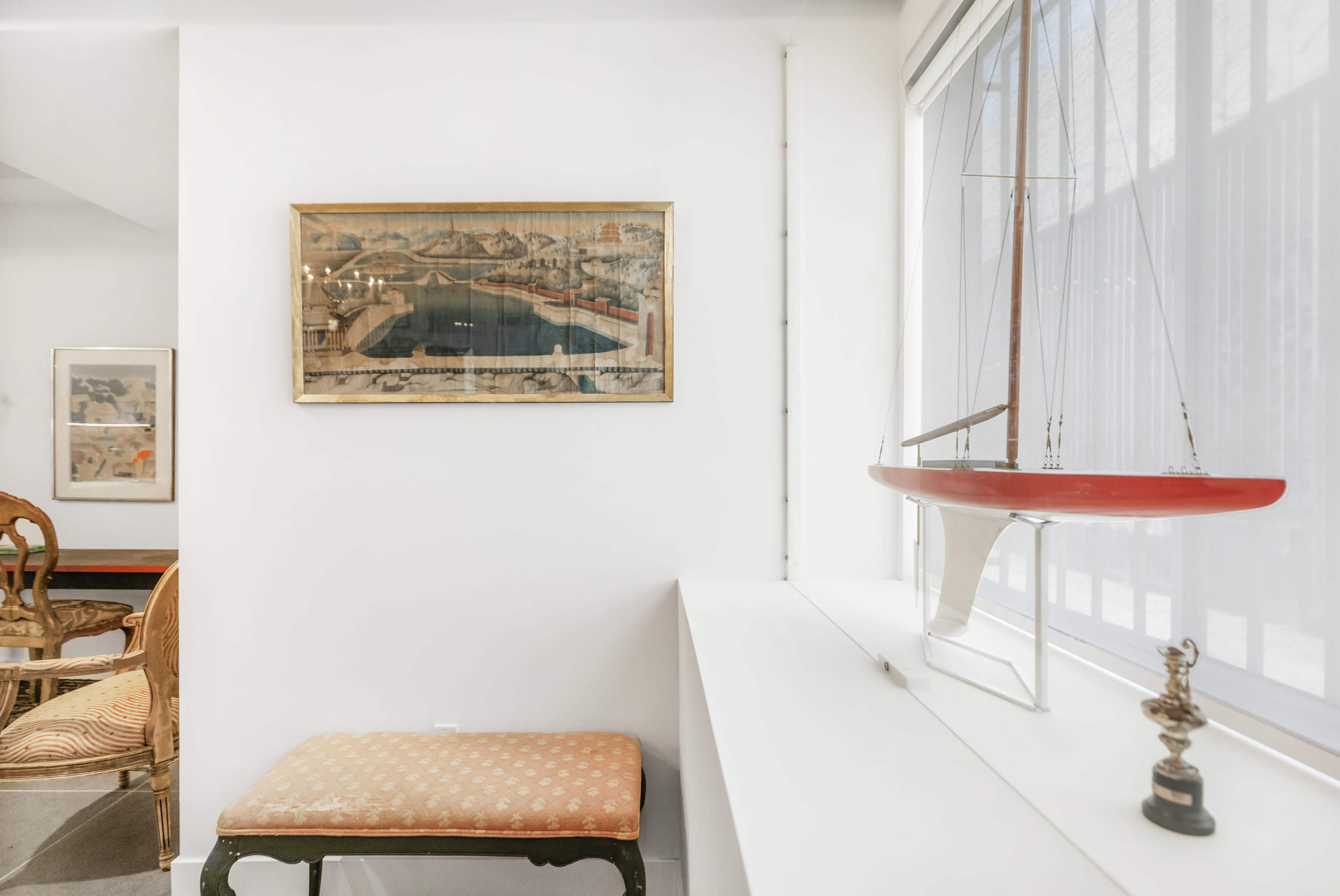 The image shows a bright, modern room featuring a framed artwork on the wall, a small boat model on a ledge, and a patterned bench with a wooden frame.