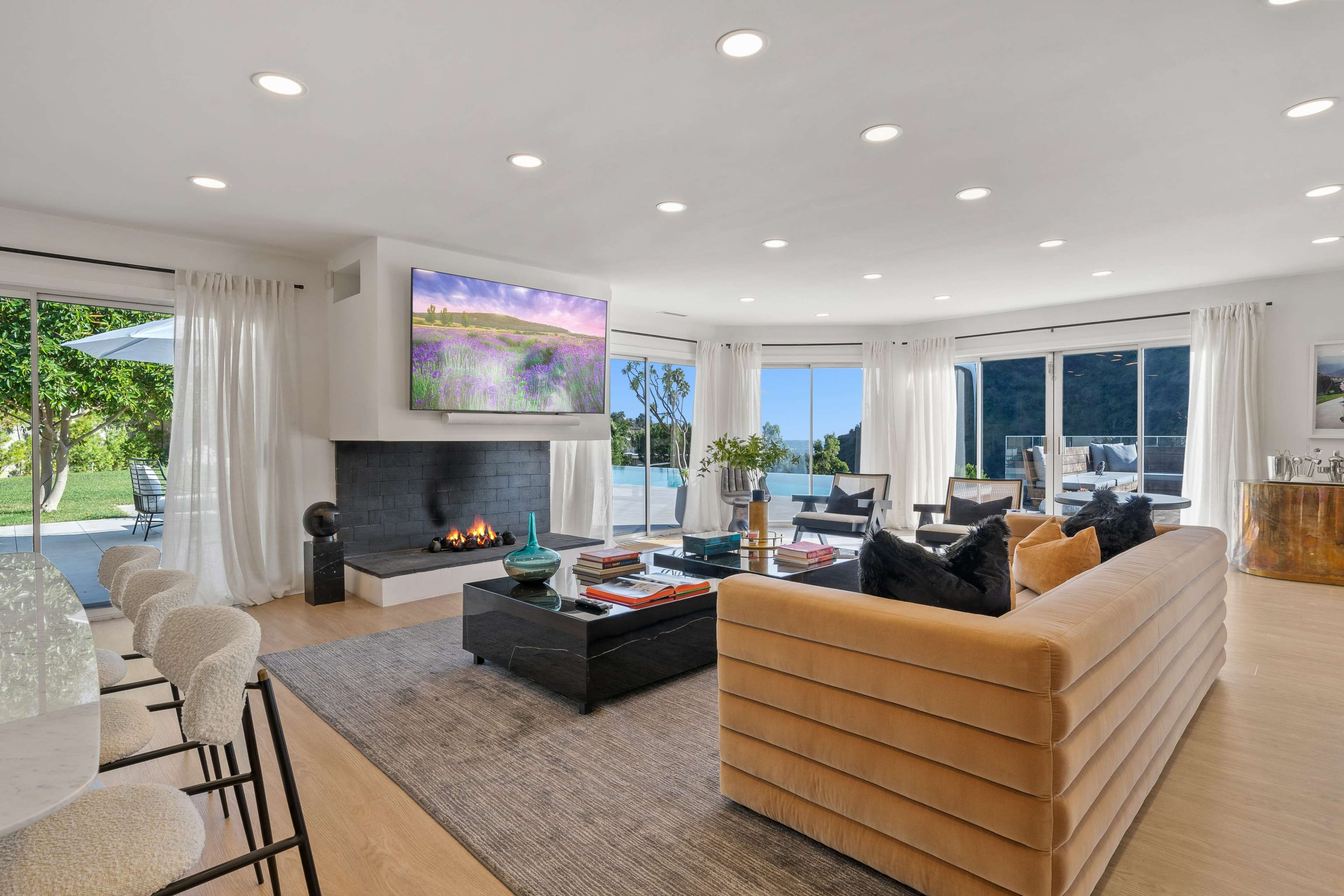 A modern living room features a beige sectional sofa, a wall-mounted TV displaying a landscape, and a large window overlooking a pool area.