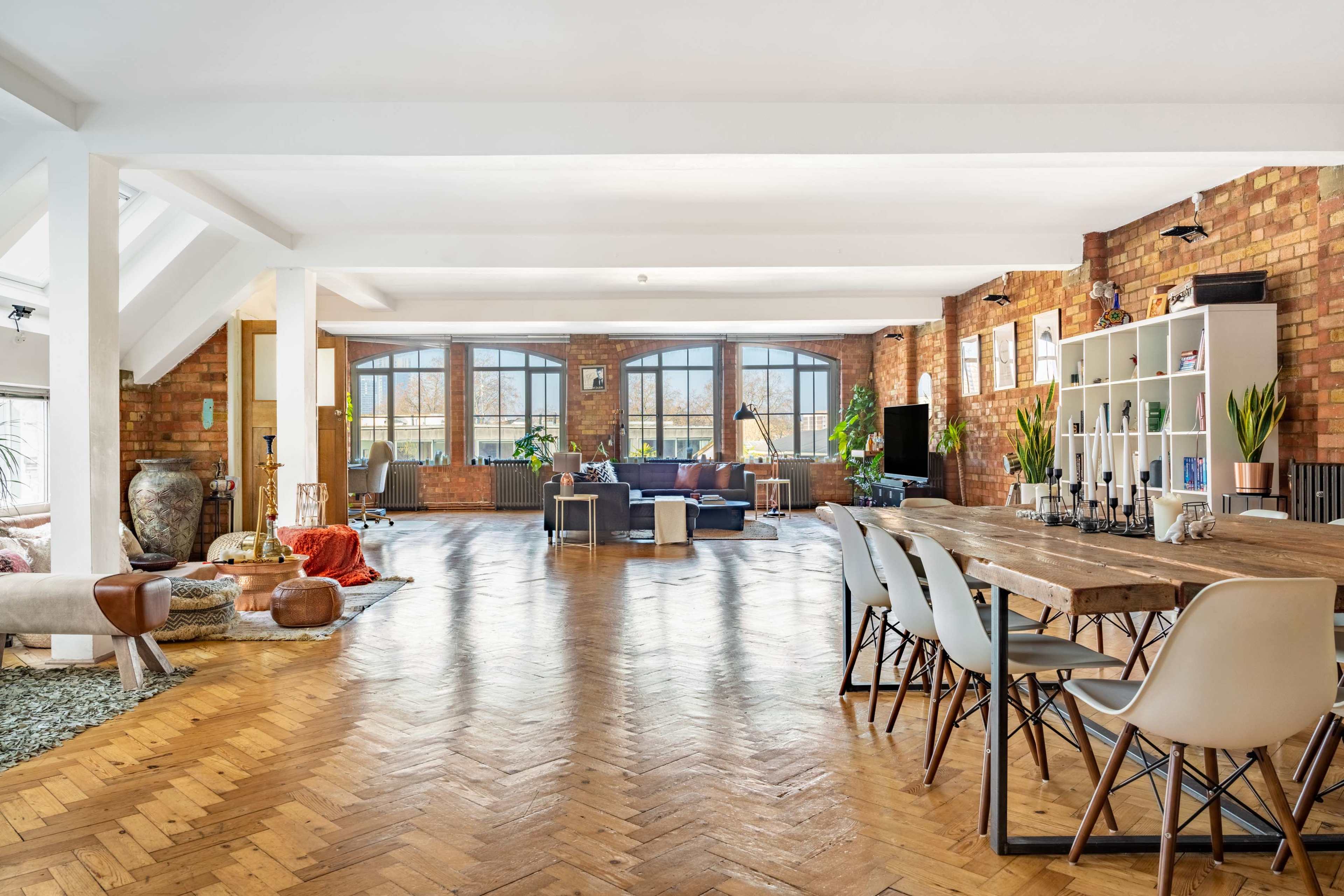 The spacious interior features exposed brick walls, large windows, and a mixture of modern furnishings and rustic decor, with a dining table in the foreground.