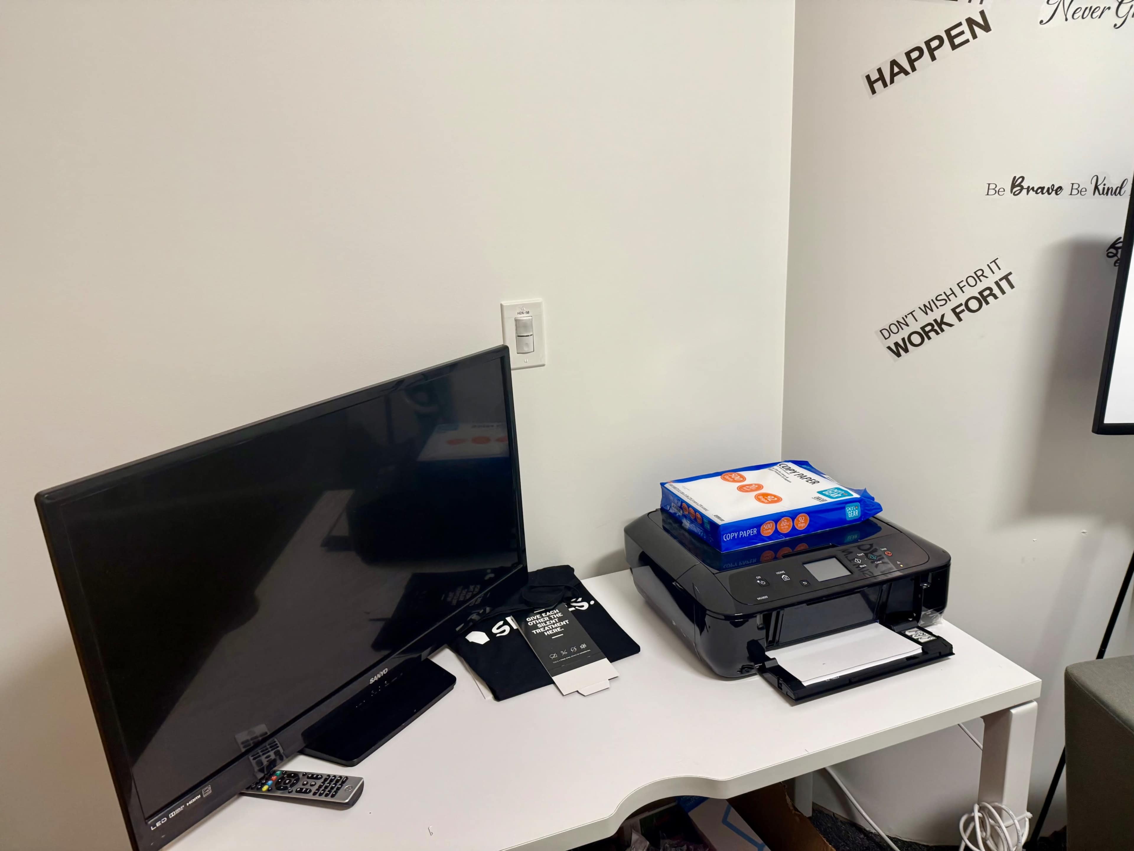 A television and a printer are placed on a white desk, accompanied by a stack of papers and a remote control.
