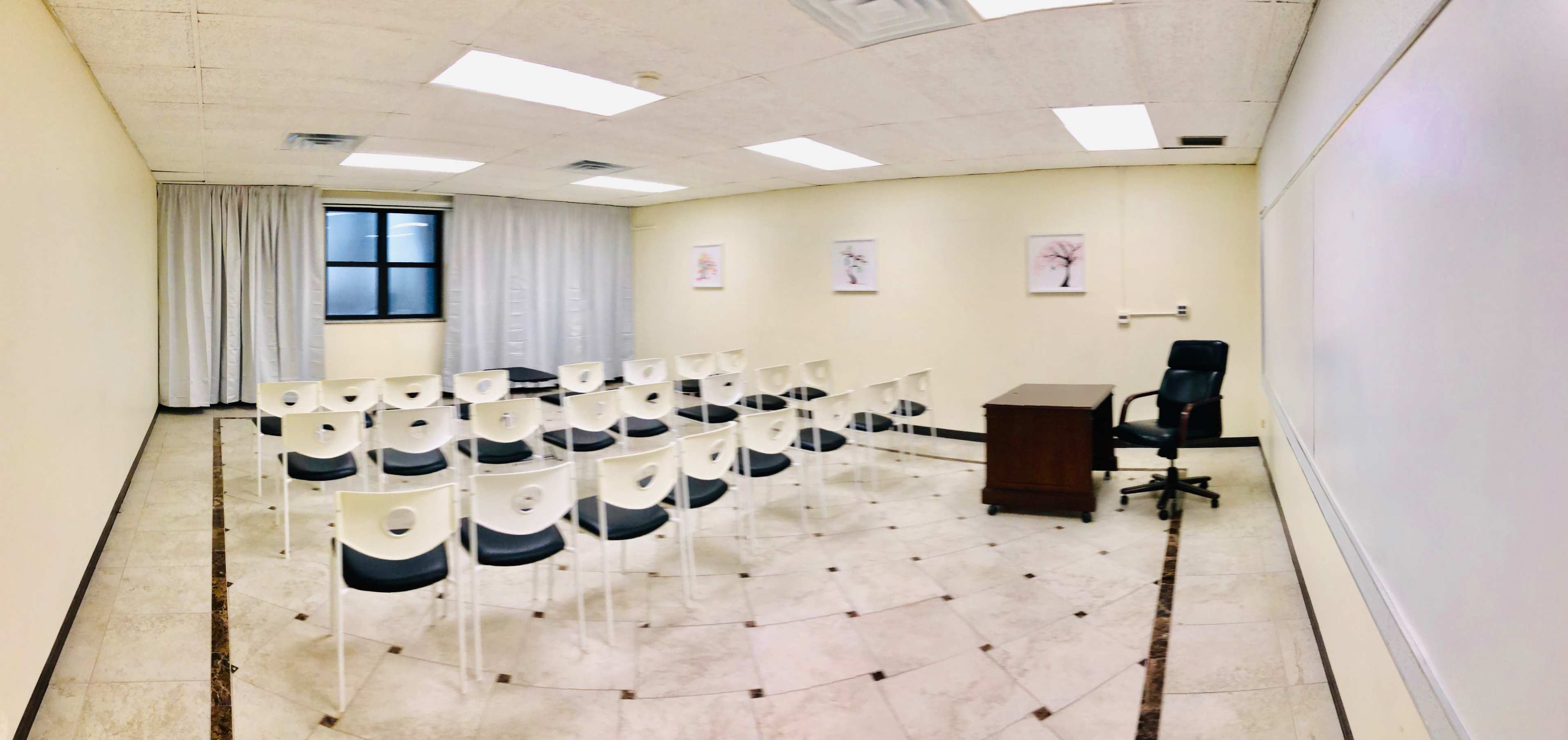 A minimalist classroom features rows of stacked white chairs with a wooden desk and a black chair at the front, illuminated by overhead lights.