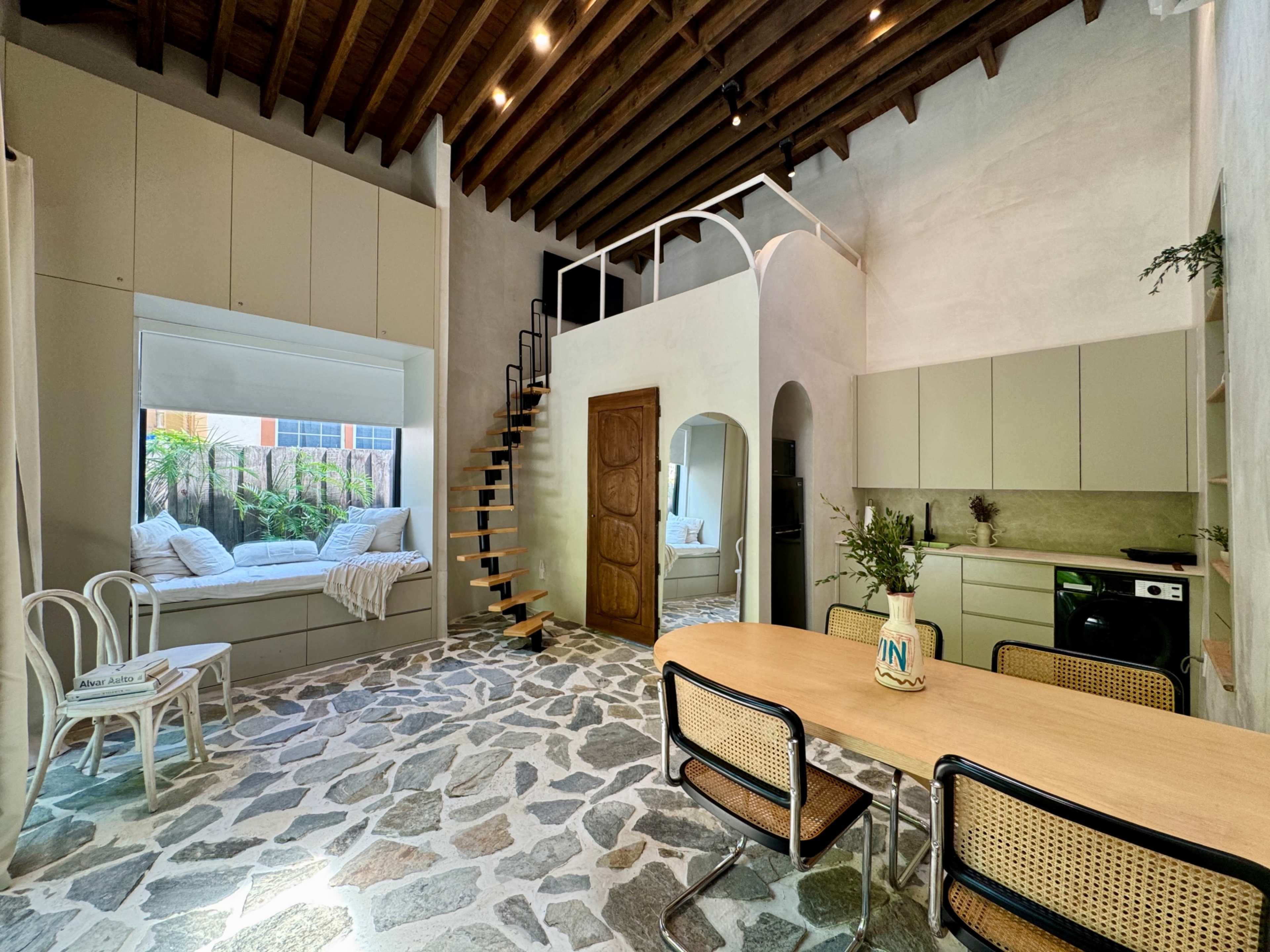 The image shows a modern interior with a stone floor, a dining area with a wooden table and chairs, a kitchenette, and a staircase leading to a loft space.
