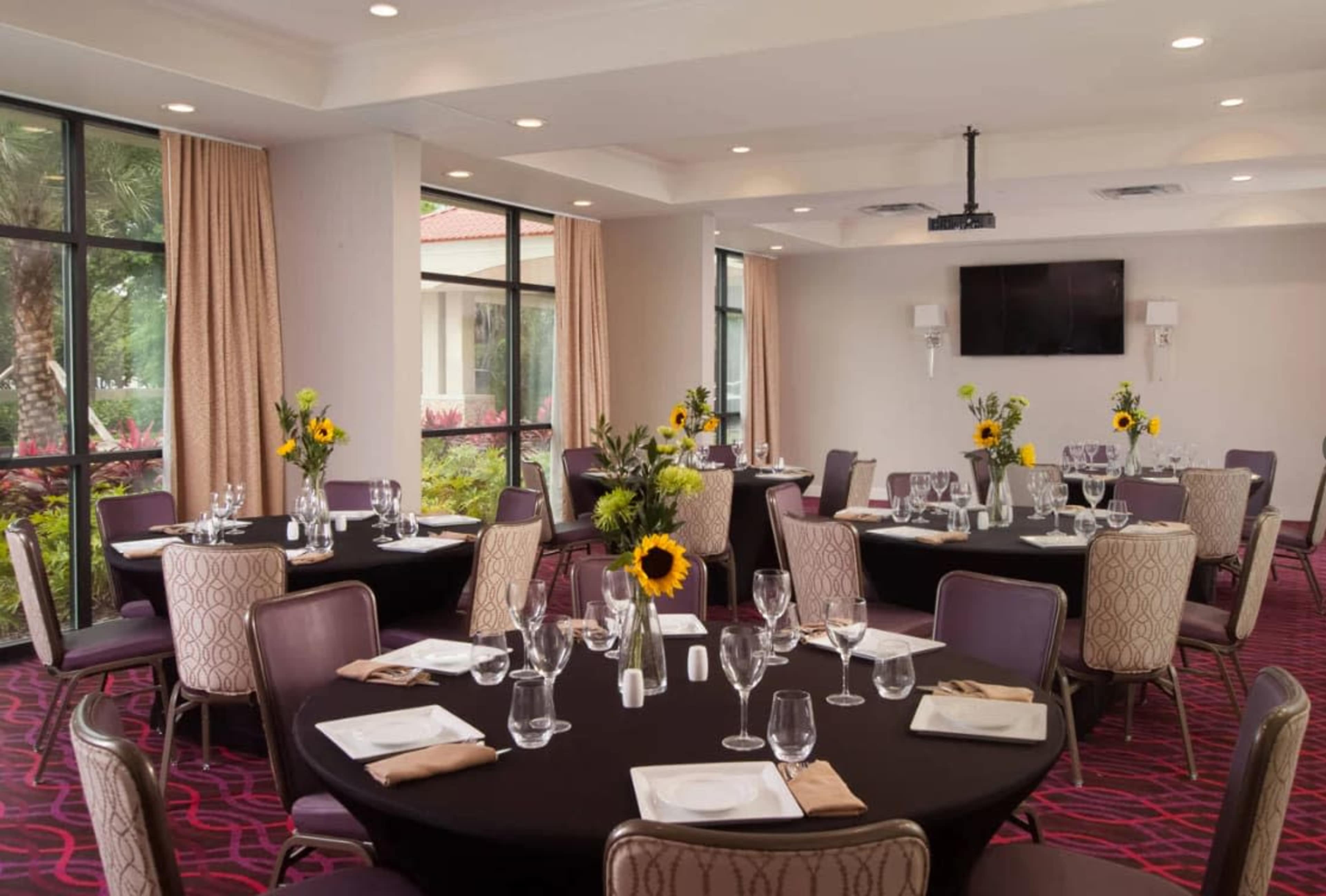 The image shows a well-lit dining room with round tables set for a meal, featuring black tablecloths and centerpieces of sunflowers.