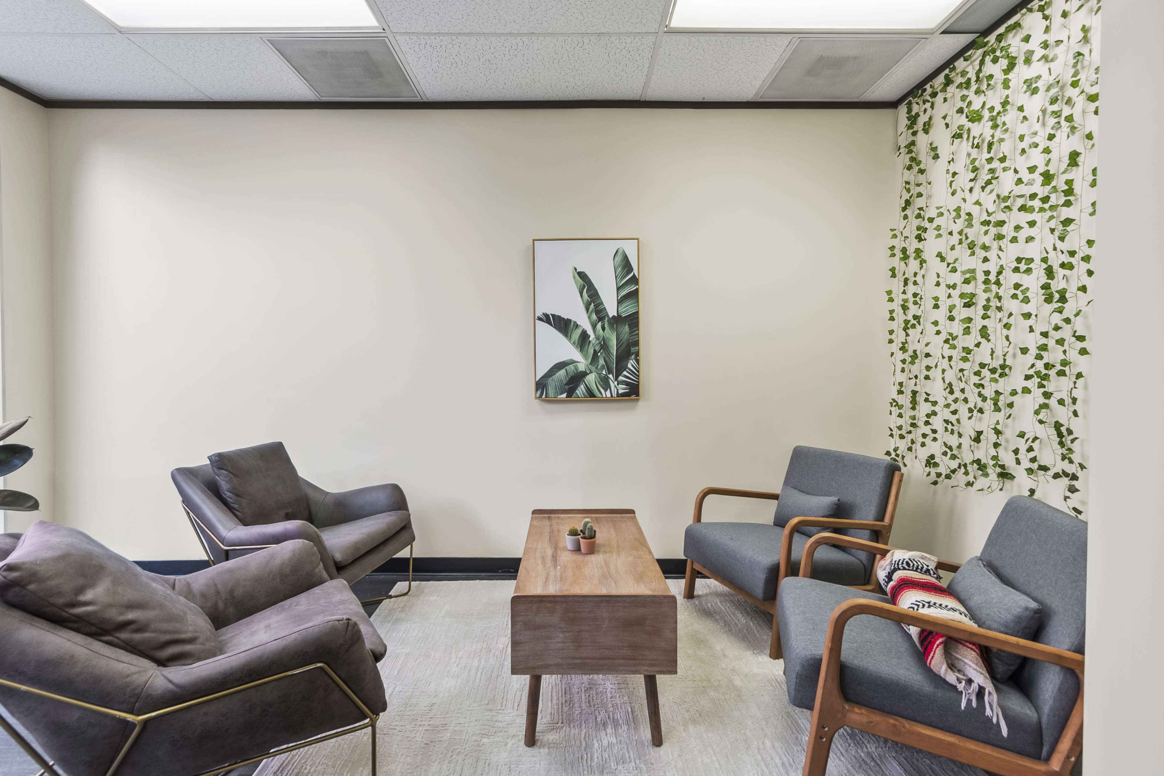 A small waiting room features two armchairs and a coffee table, with a plant-themed artwork on the wall and greenery near the window.