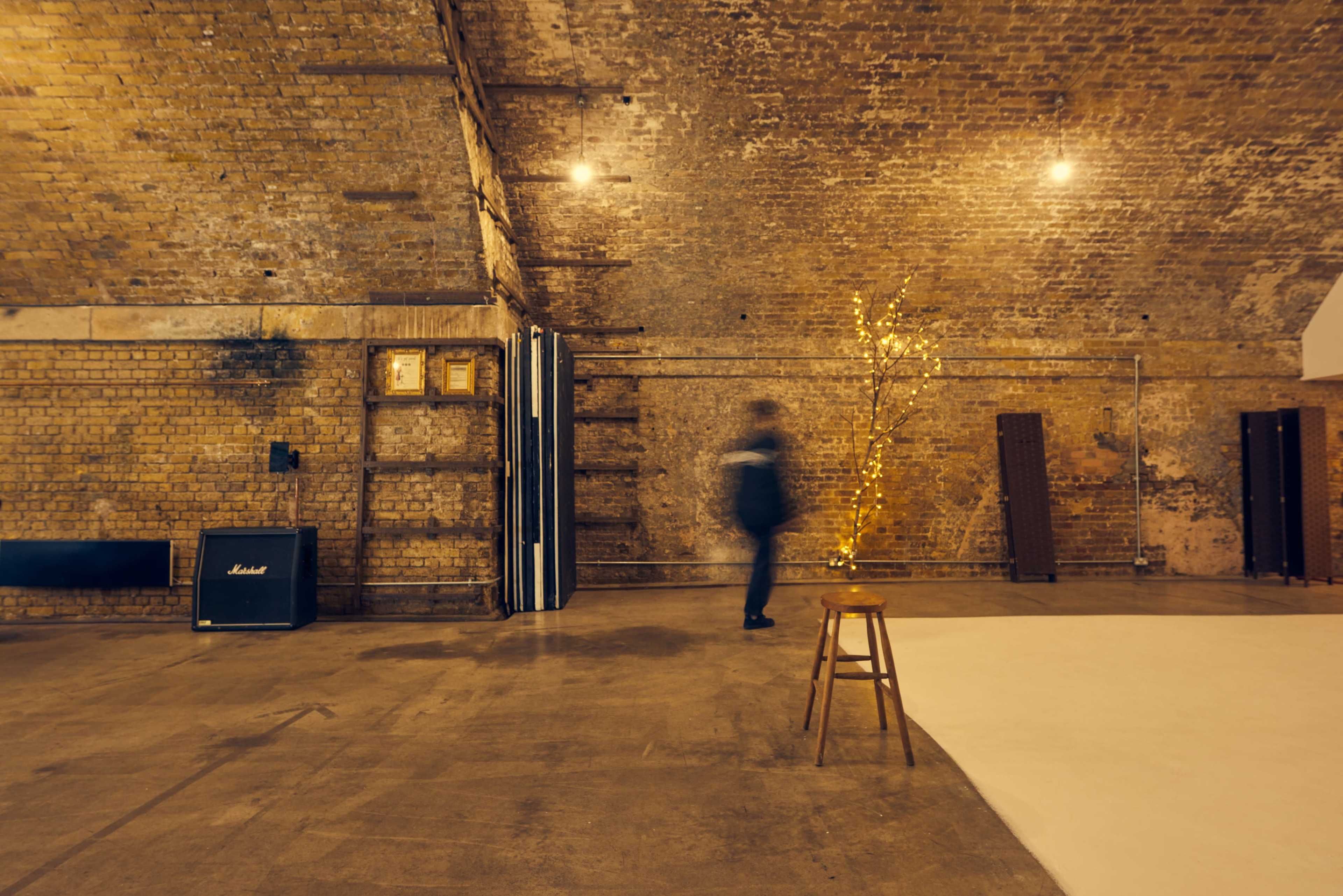 A person walks past a stool and a small, illuminated tree in a spacious, brick-walled interior.