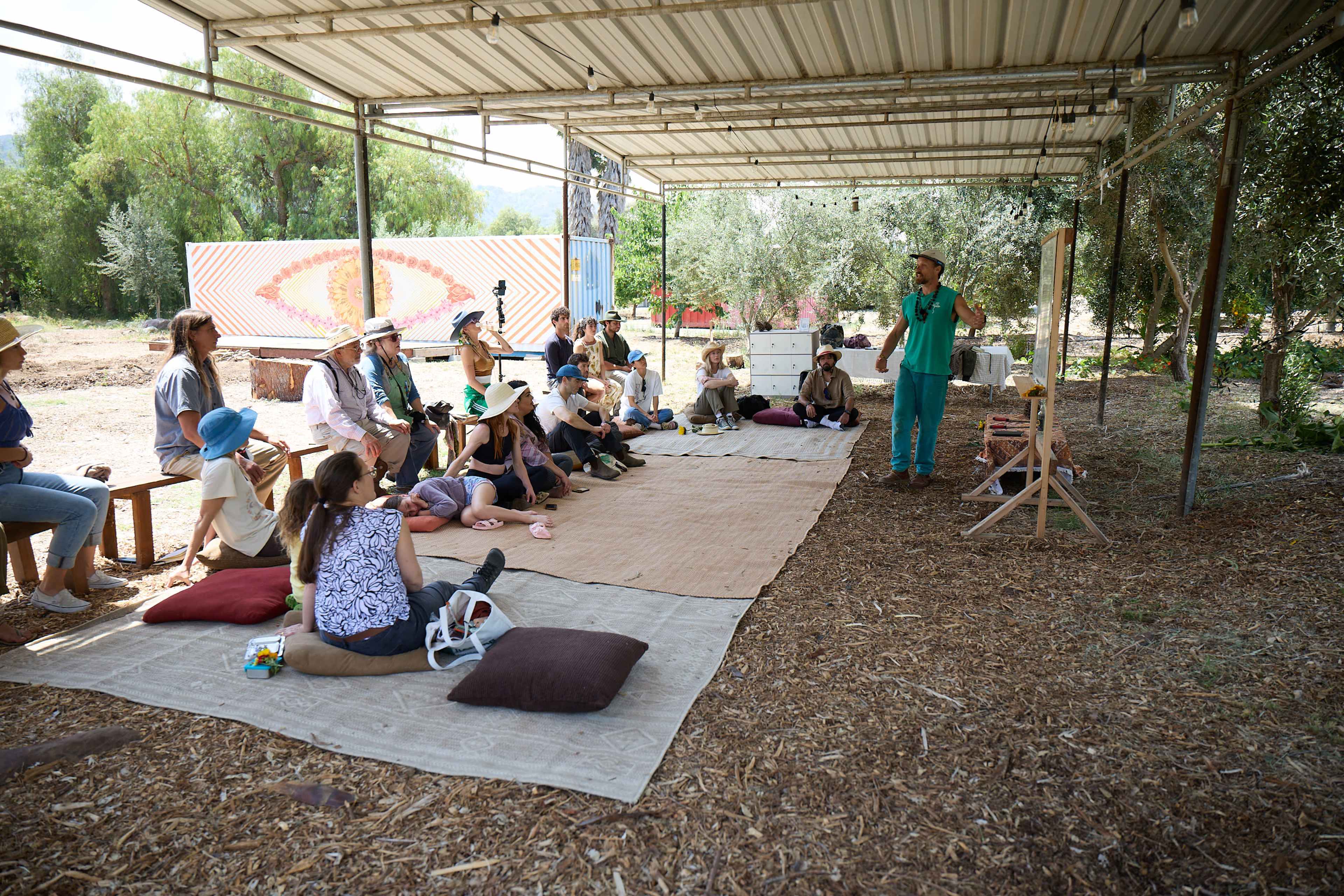 A group of people sits on mats under a shaded structure while a speaker addresses them in an outdoor setting.