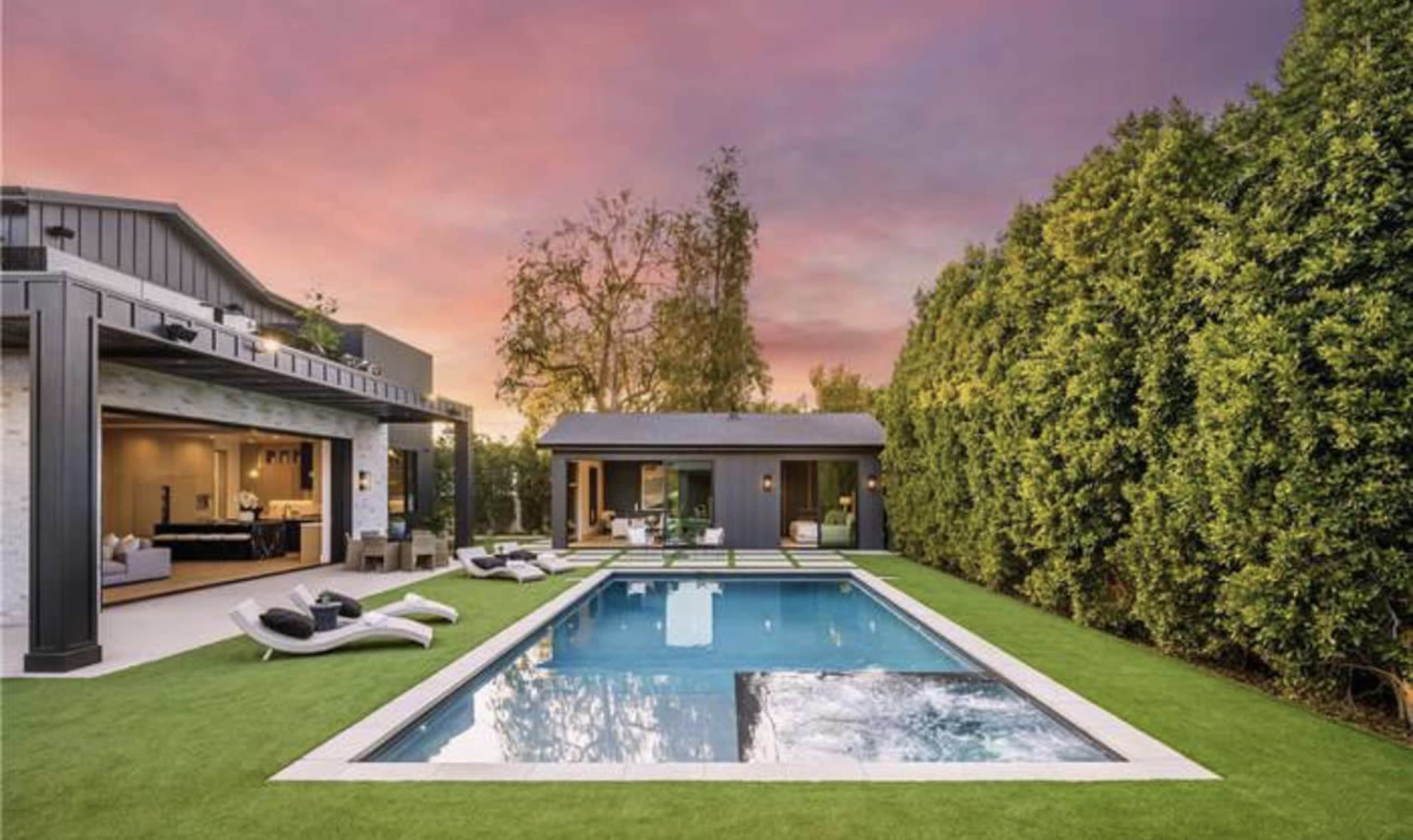 A modern backyard featuring a rectangular swimming pool surrounded by landscaped grass and lounge chairs, with a stylish home in the background against a colorful sunset sky.