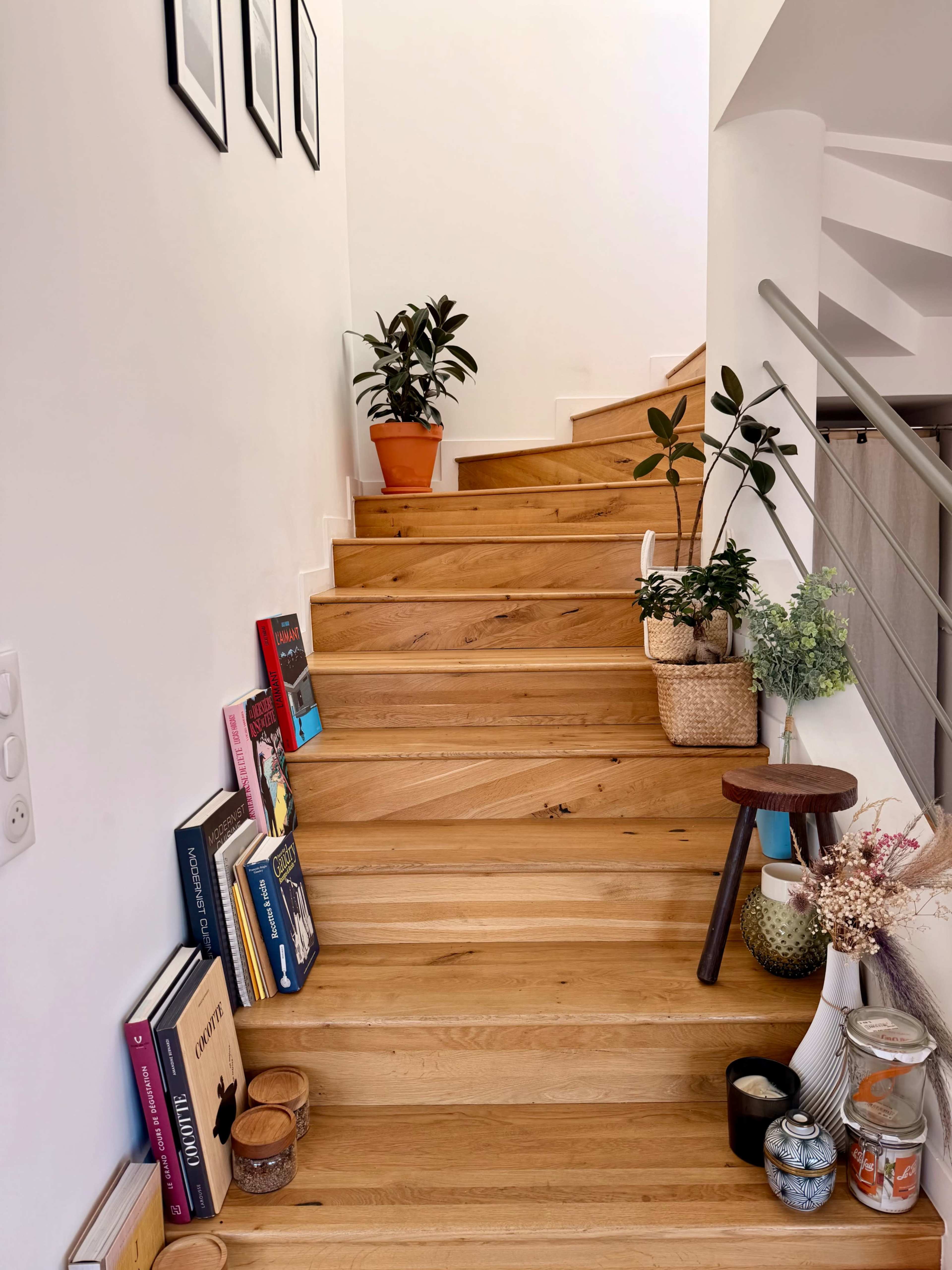The image shows a wooden staircase with potted plants and books placed on the steps, along with decorative items and a small stool at the base.