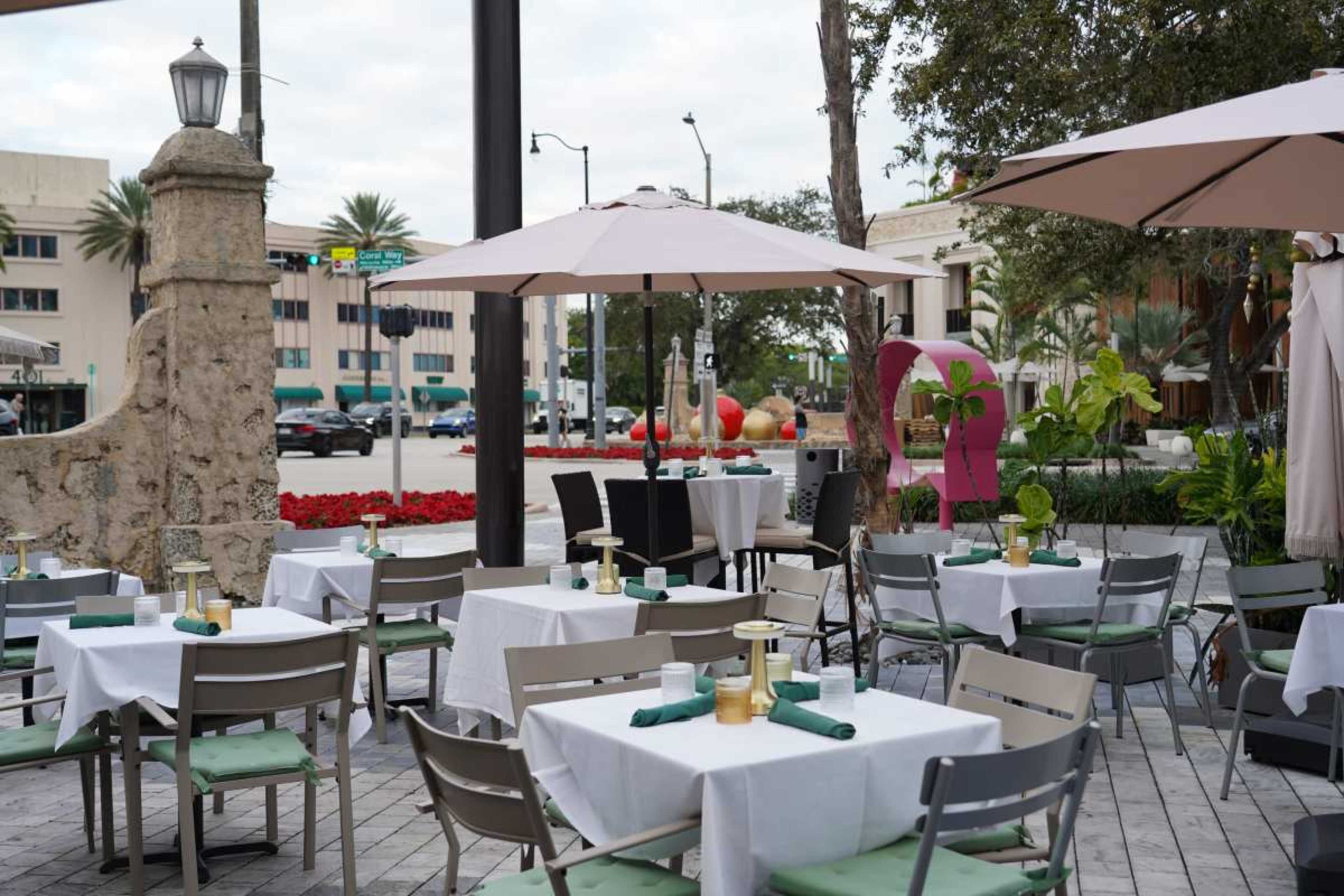 The image shows an outdoor restaurant seating area with tables covered in white linens and umbrellas, surrounded by plants and a street view in the background.