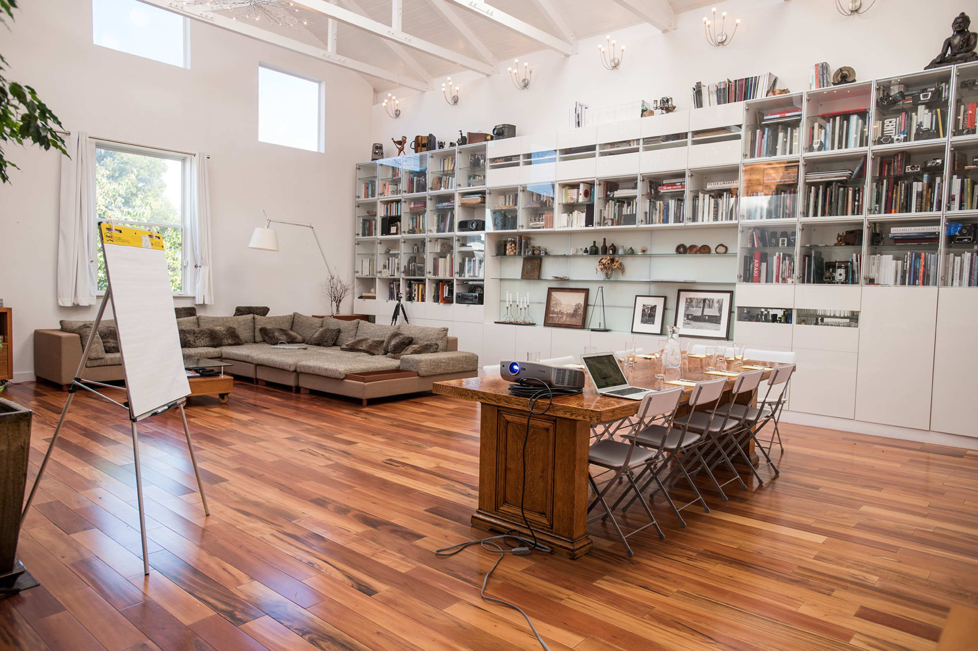 The image shows a modern meeting room with a wooden table set for a presentation, chairs arranged around it, and a large bookshelf filled with books along one wall.