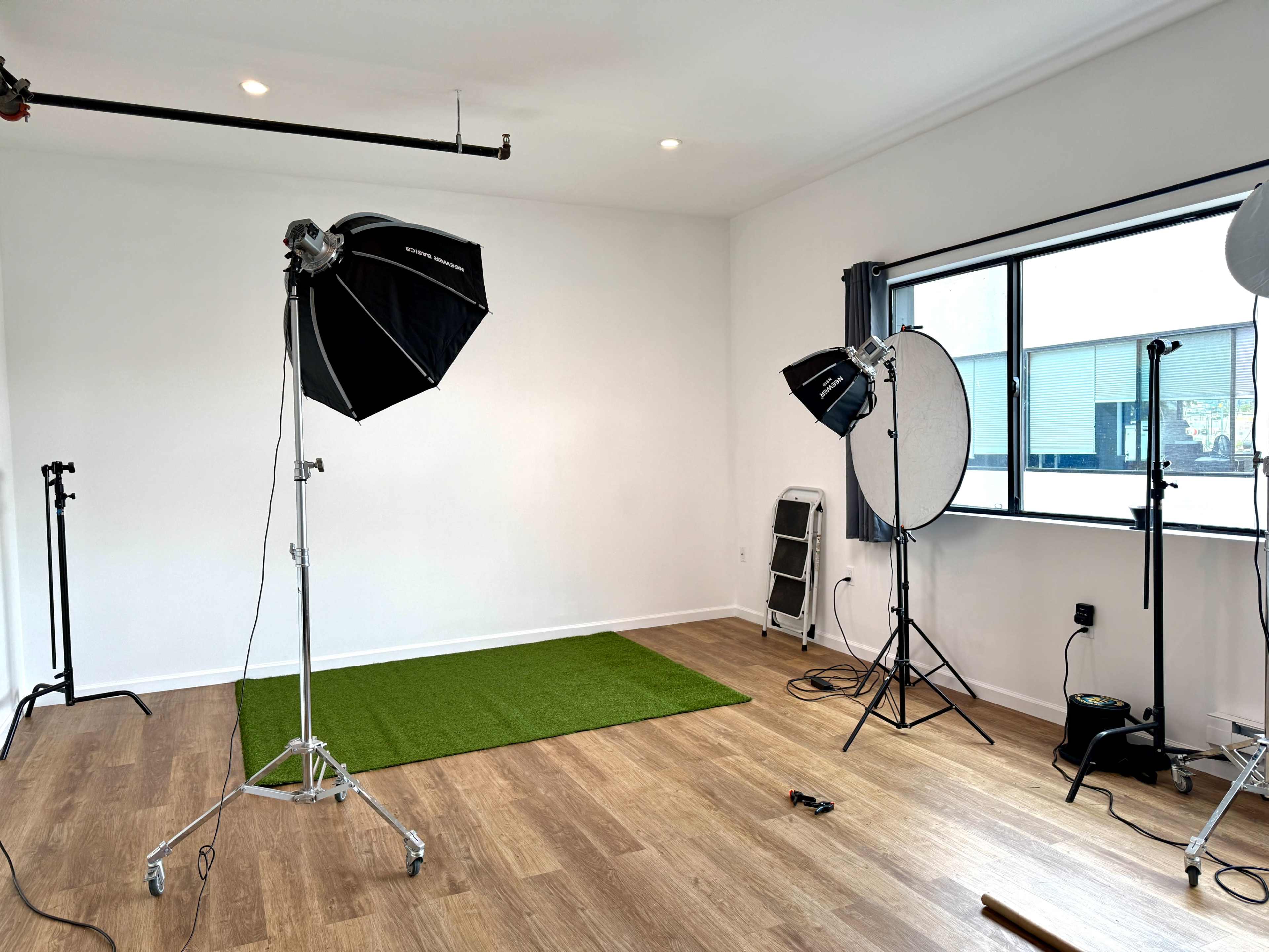 The image shows a photography studio with two lighting setups, a green carpet area, and a large window.