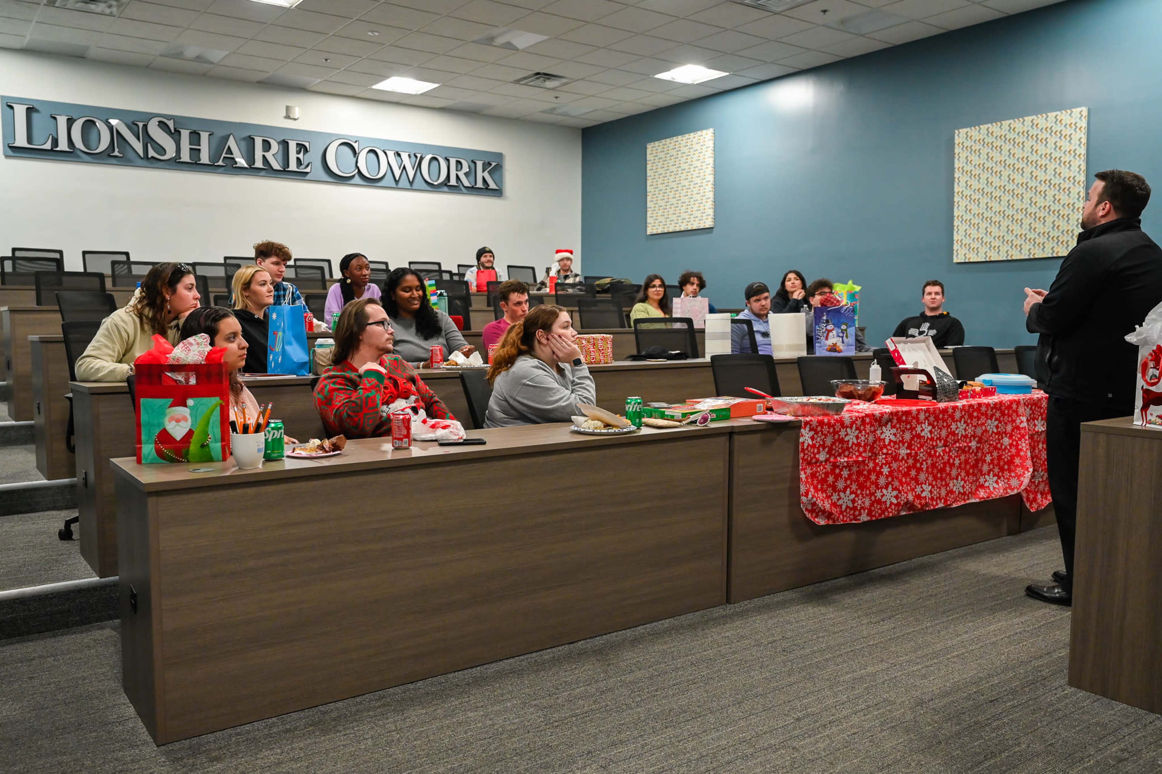 A group of people is gathered in a meeting room at LionShare Cowork, with festive decorations and gifts on several tables.
