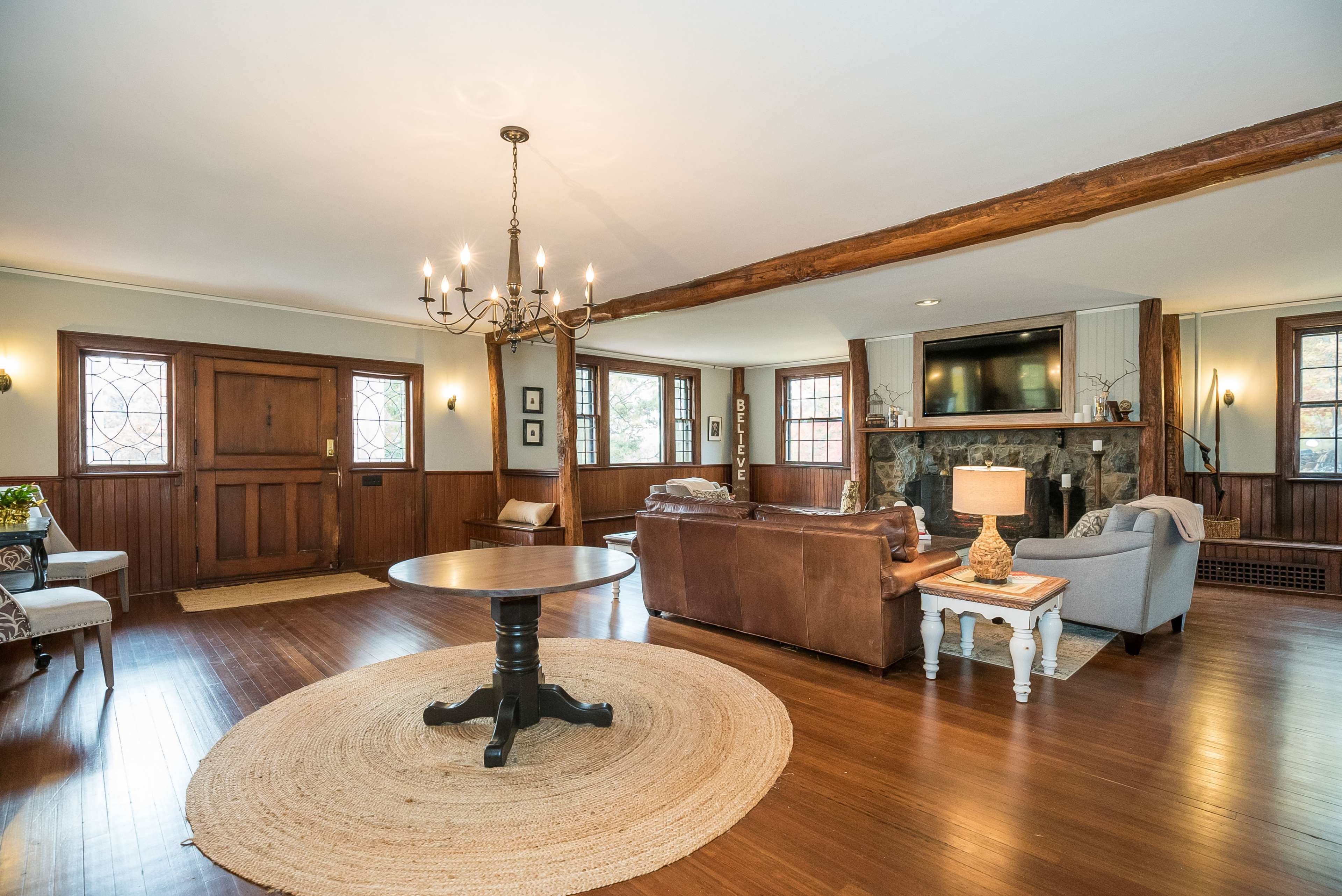 A spacious living room featuring a wooden floor, a central circular table, a fireplace, and large windows with decorative panes.