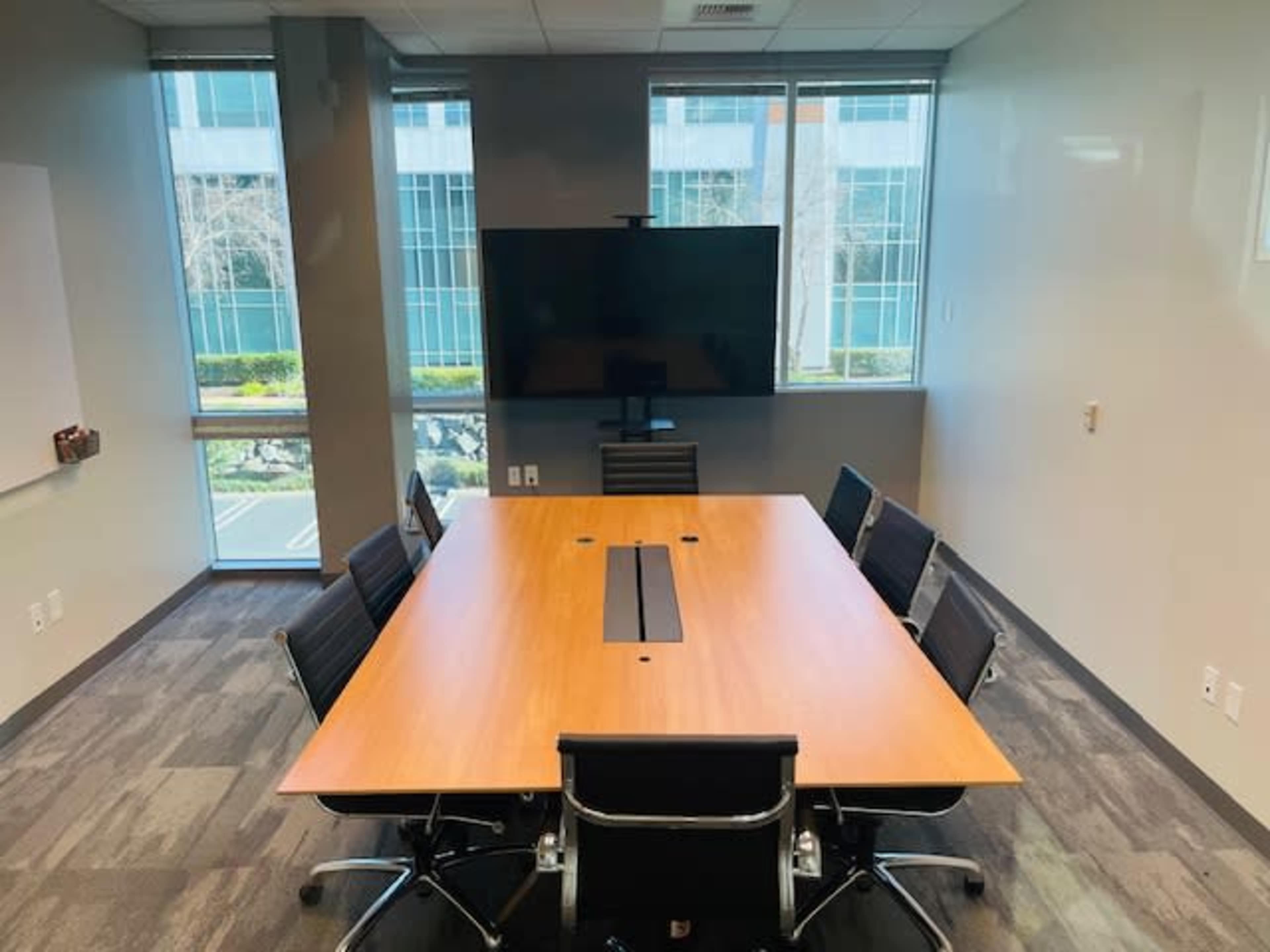 A modern conference room features a large wooden table with eight black chairs and a wall-mounted screen.