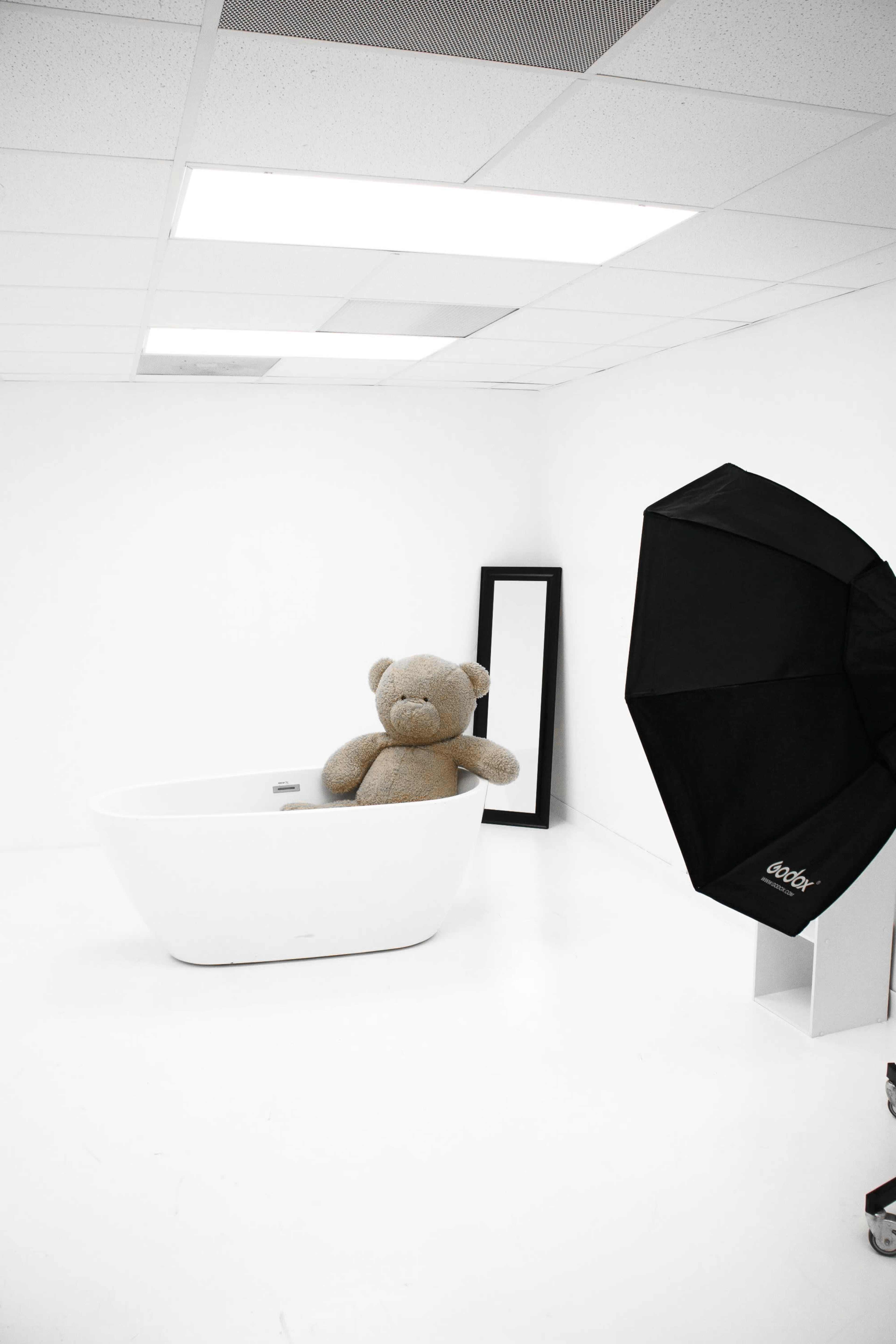 A large teddy bear sits in a white bathtub in a minimally decorated studio with bright lighting and a mirror.