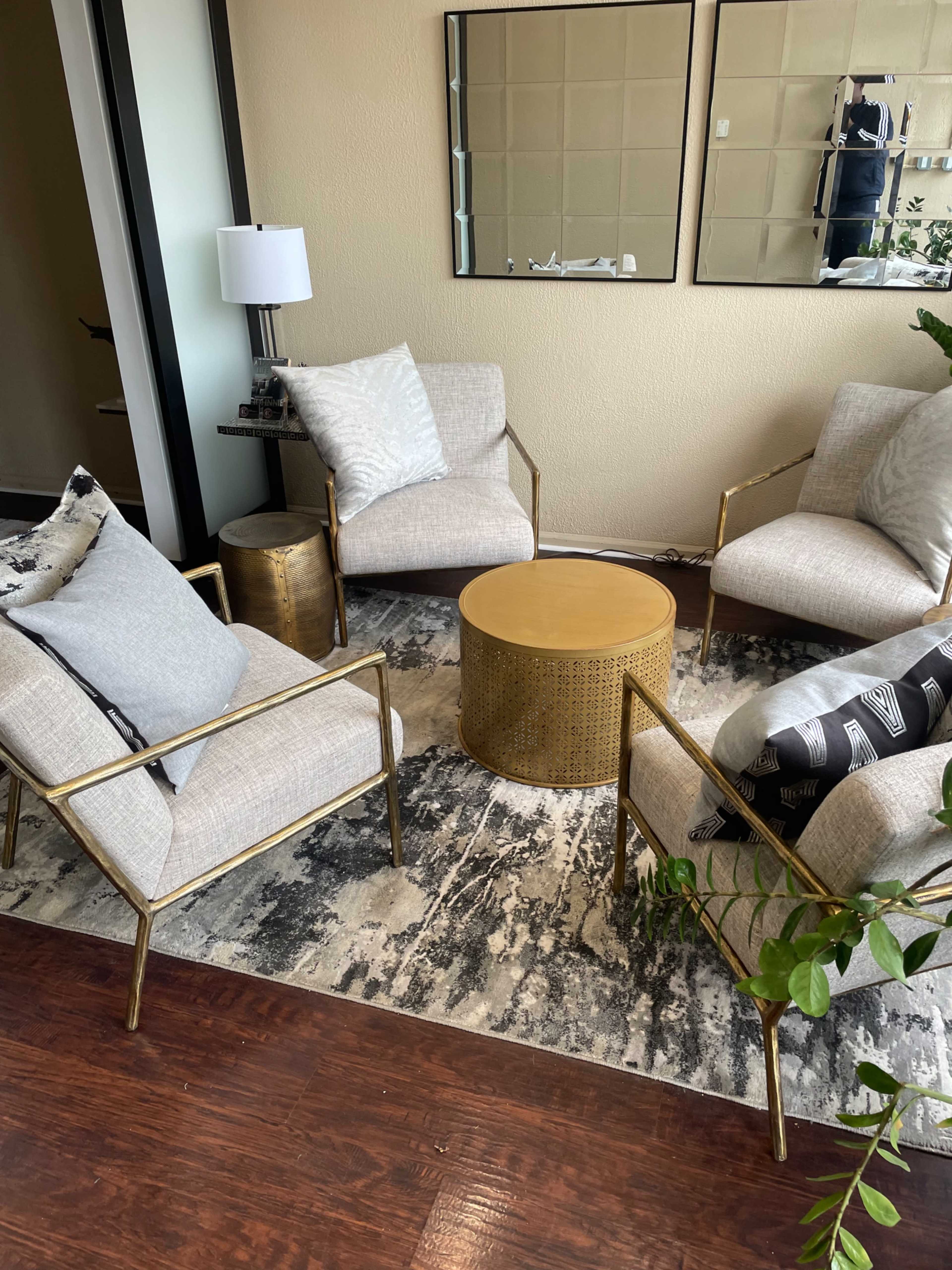 A modern seating arrangement features two light-colored chairs with cushions, a round gold coffee table, and a patterned rug in a well-lit room.