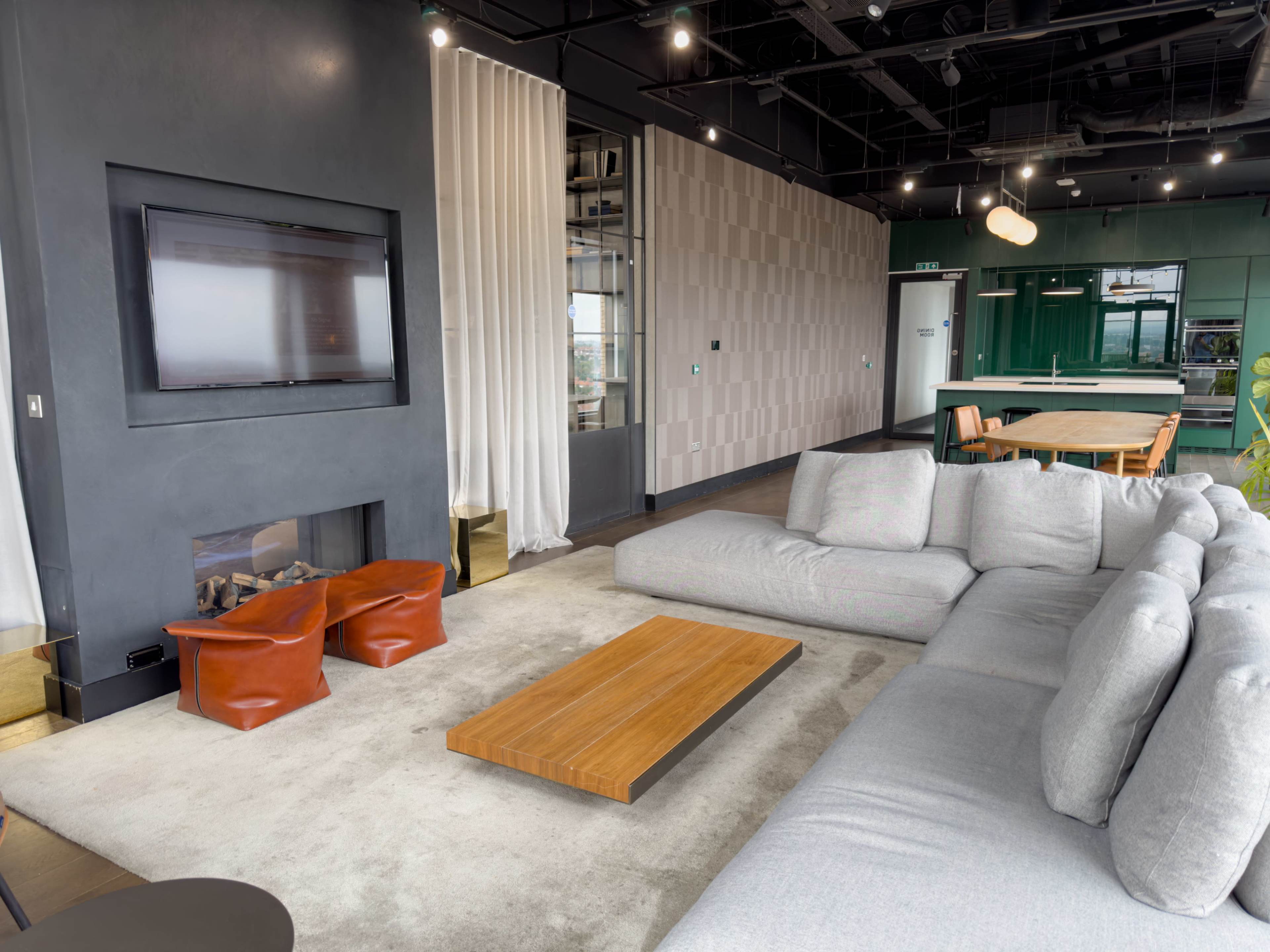 The image shows a modern lounge area featuring a gray sectional sofa, a wooden coffee table, and two small brown stools, with a flat-screen TV mounted on the wall and a green kitchen area visible in the background.