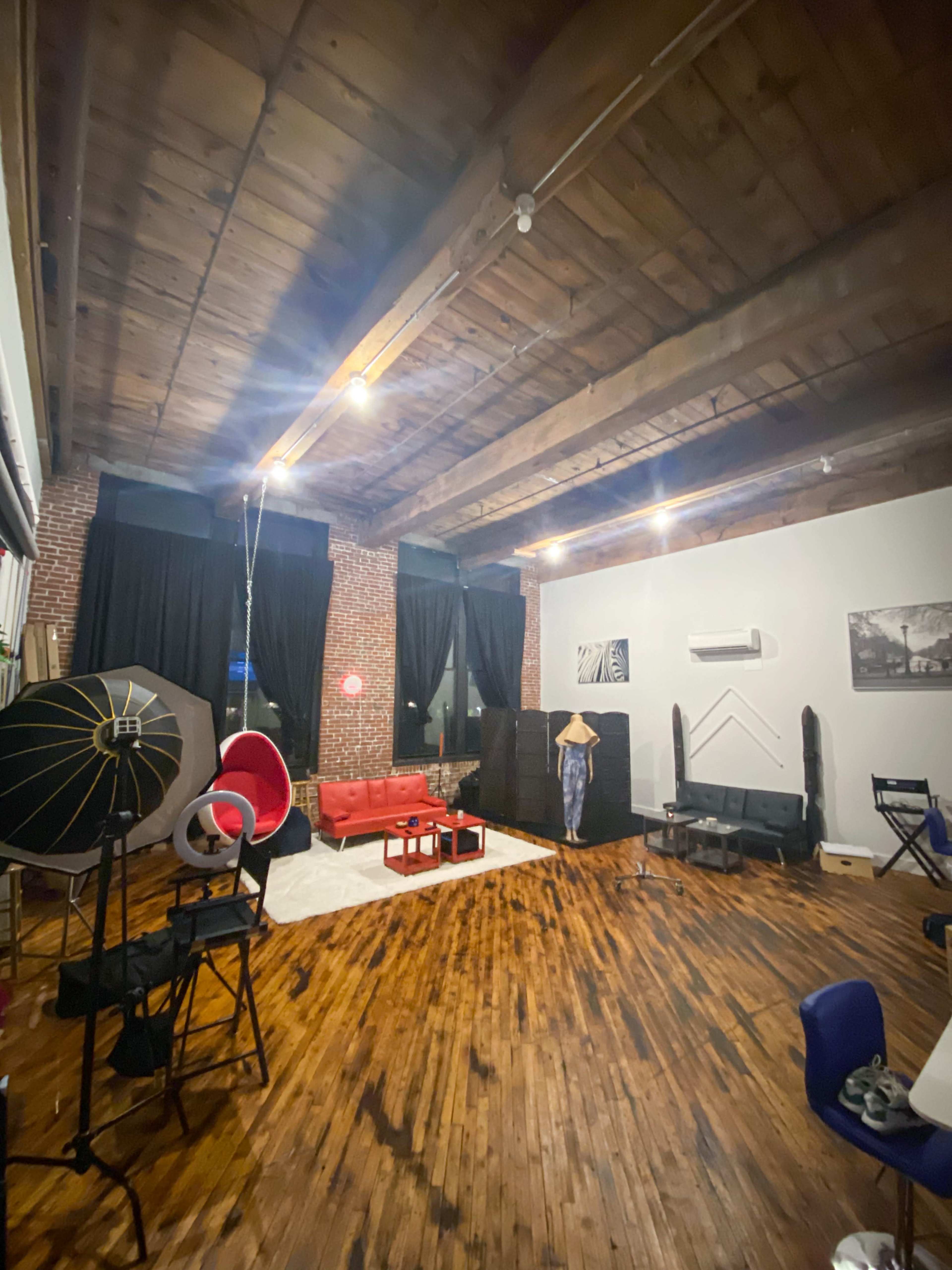 A spacious room with wooden floors, exposed brick walls, and various furniture pieces, including a red sofa, folding chairs, and a light source setup.