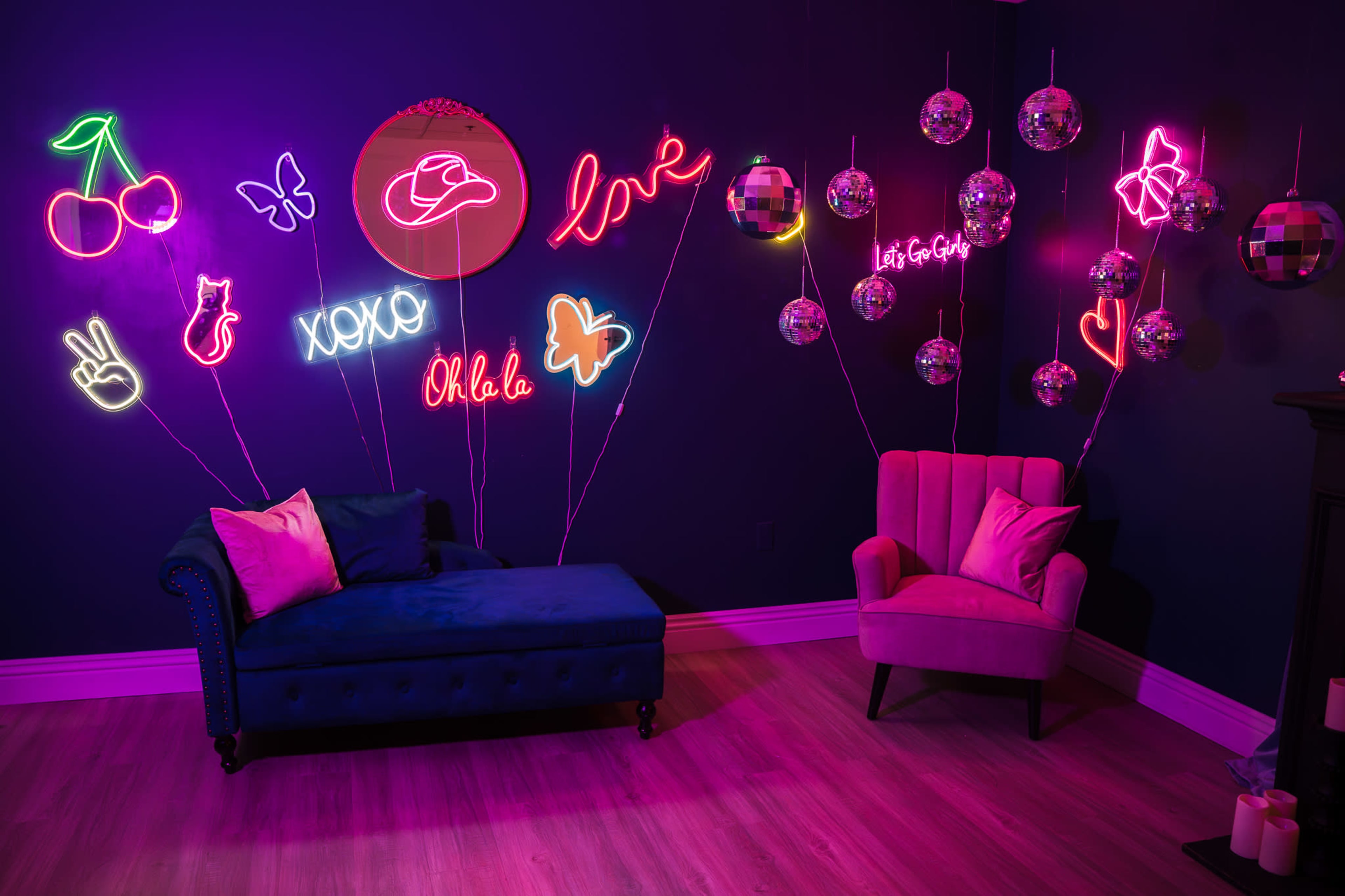The image features a brightly lit room with neon signs, a blue couch, a pink chair, and decorative disco balls hanging on the wall.
