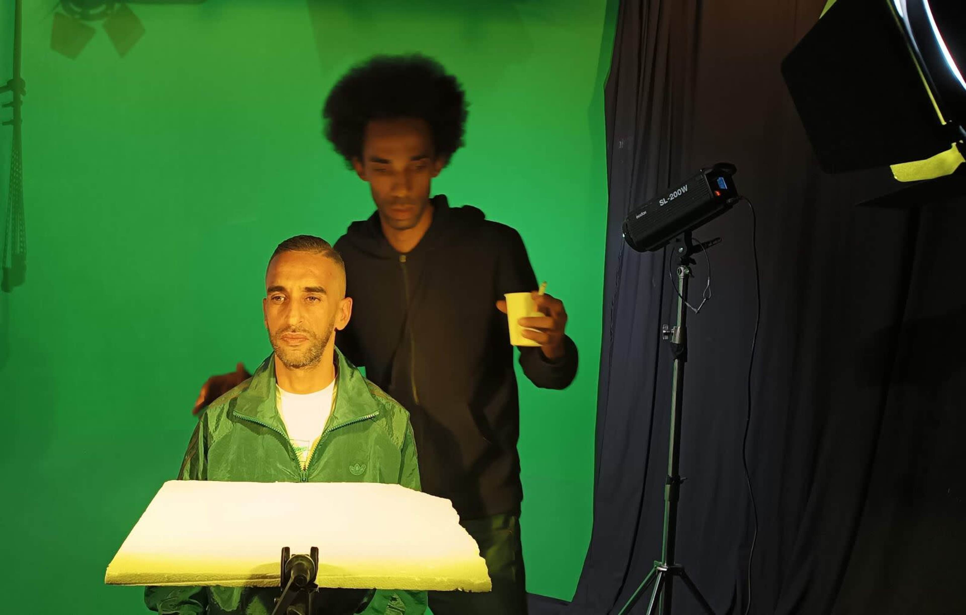A man sits in front of a green screen while another man stands behind him holding a cup.