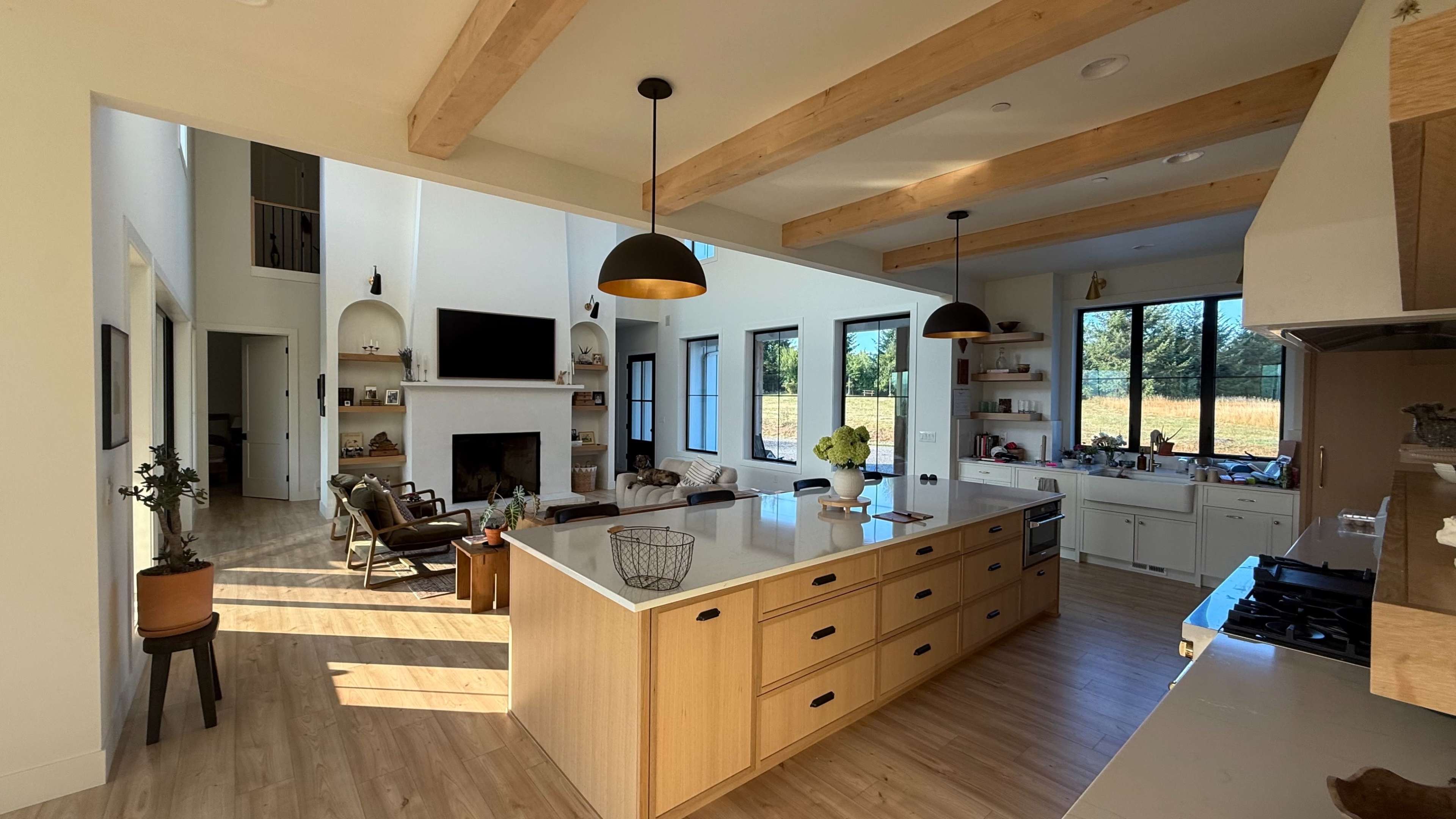The image shows a spacious, modern kitchen and living area with large windows, wooden beams, and an island countertop, featuring an open layout that connects to a bright, airy space.