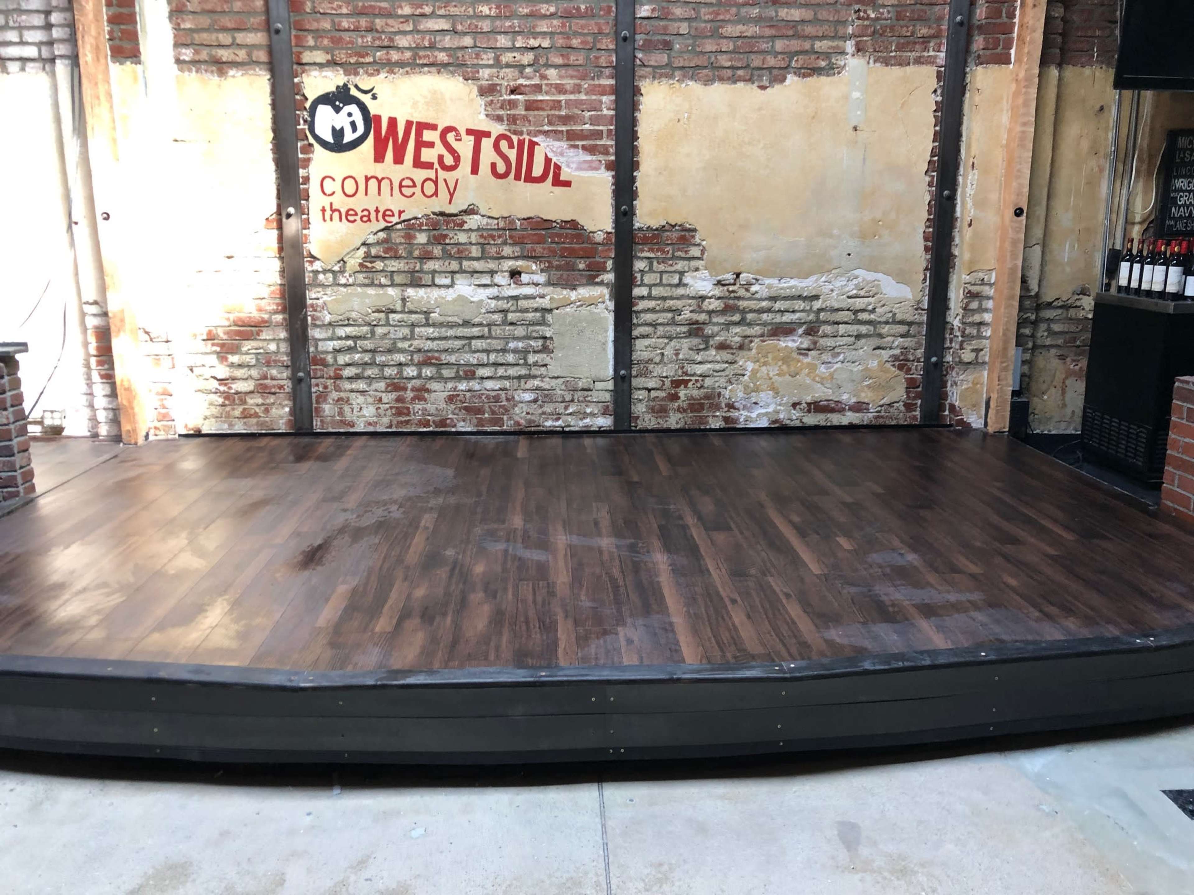 A stage area with a wooden floor, brick walls, and a sign for the Westside Comedy Theater.