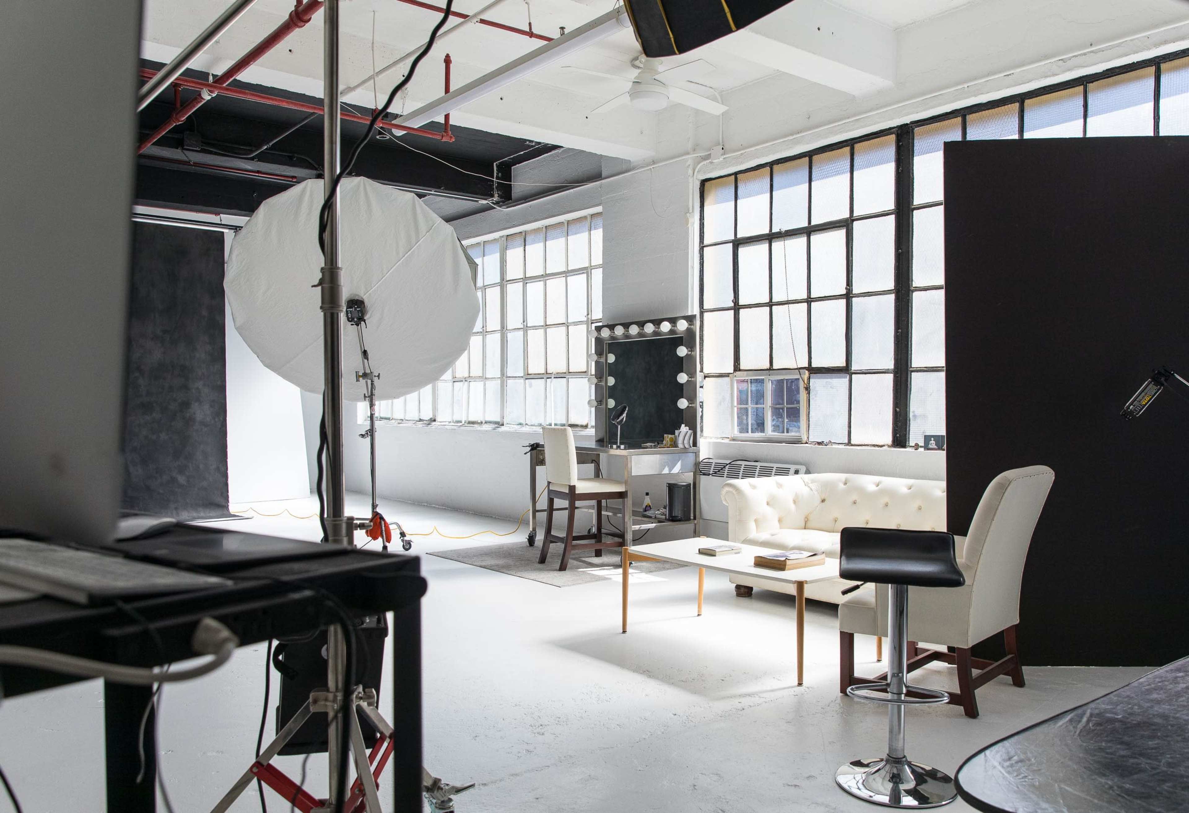 The image shows a bright photo studio with large windows, a makeup station, a sofa and chairs, and professional lighting equipment.