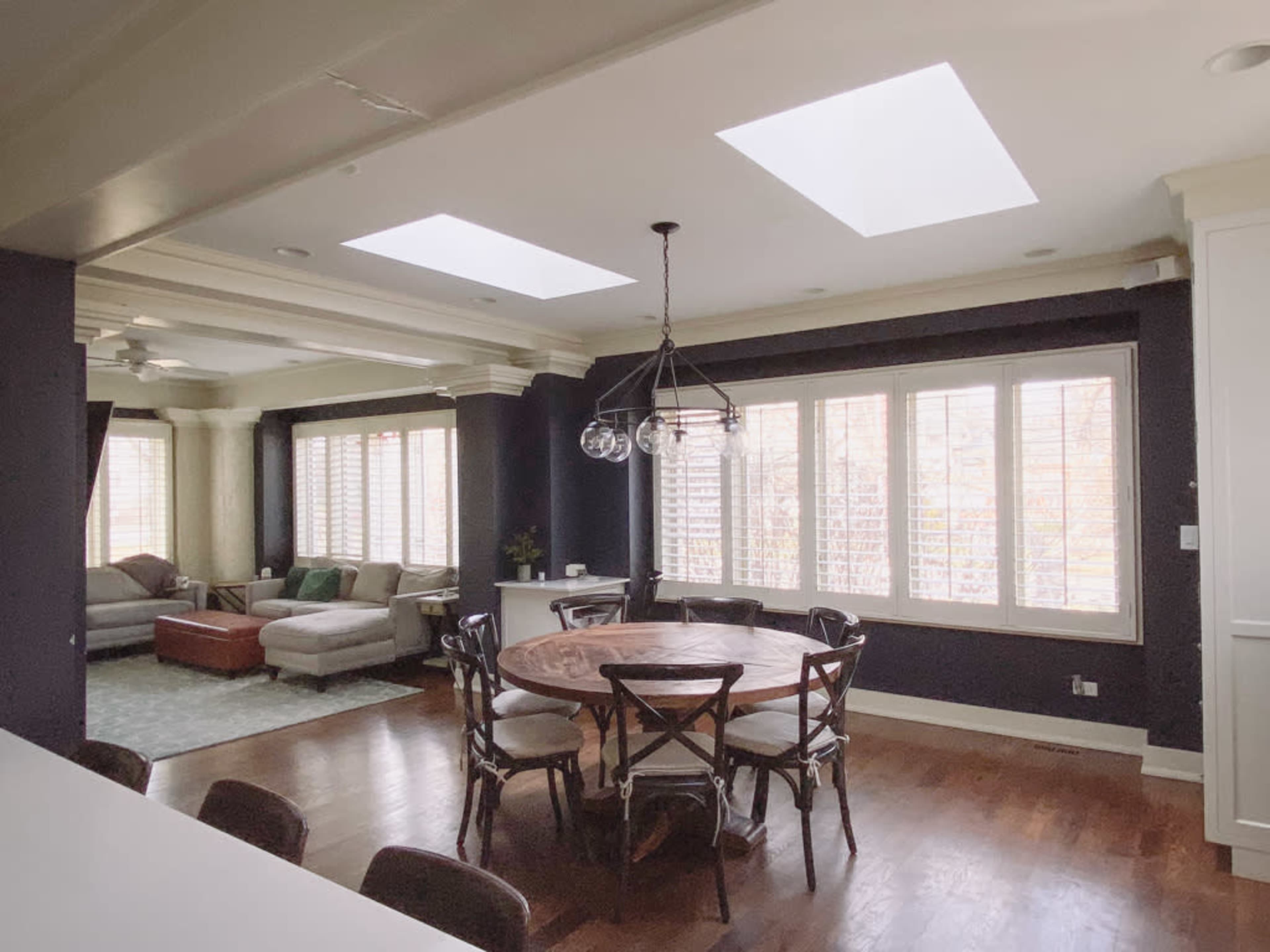 The image shows a dining area with a round wooden table surrounded by chairs, adjacent to a living space with large windows and a sectional sofa.